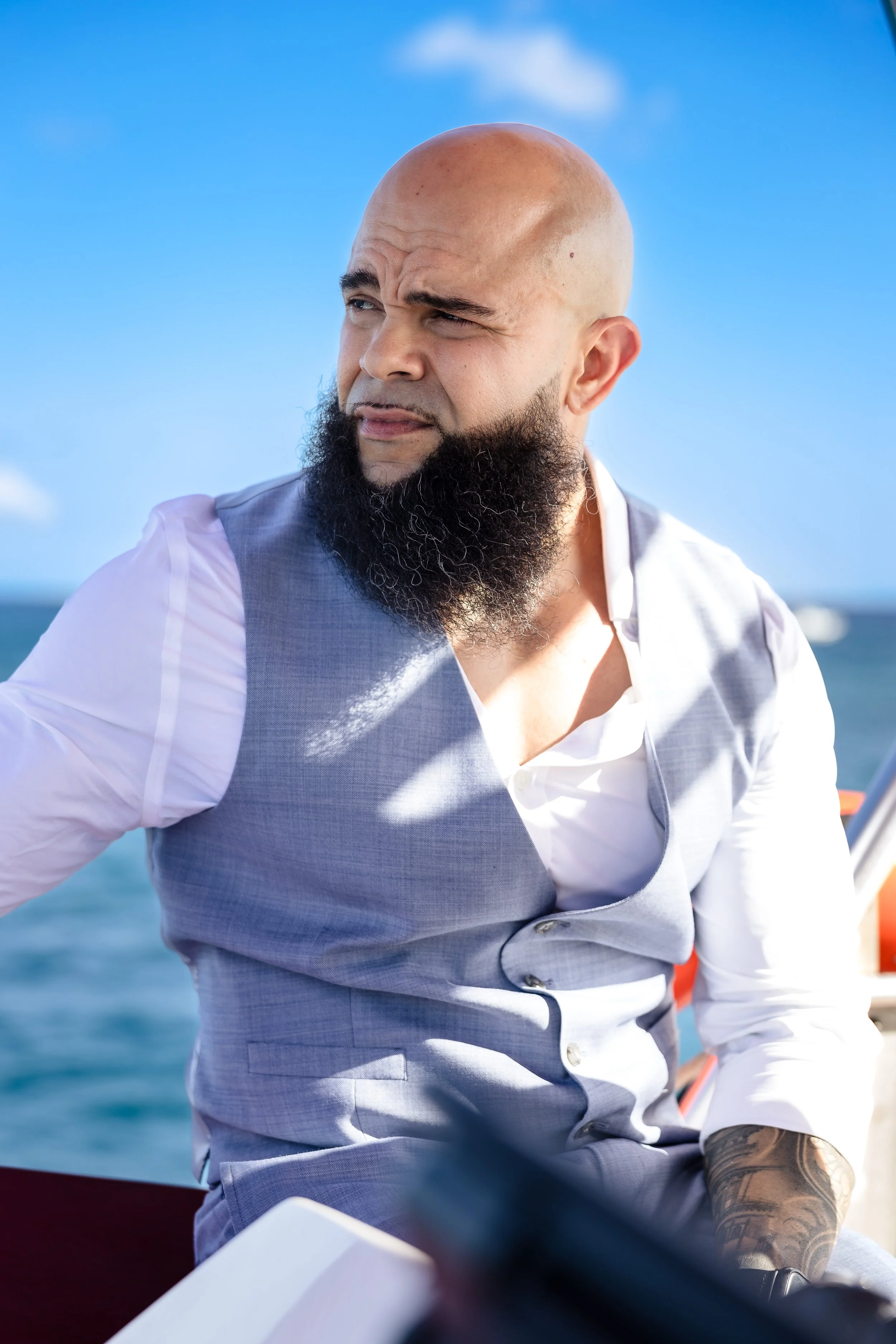 A man with a shaved head and a beard wearing a light gray vest and white shirt, looking to his left, on a boat with the ocean in the background.