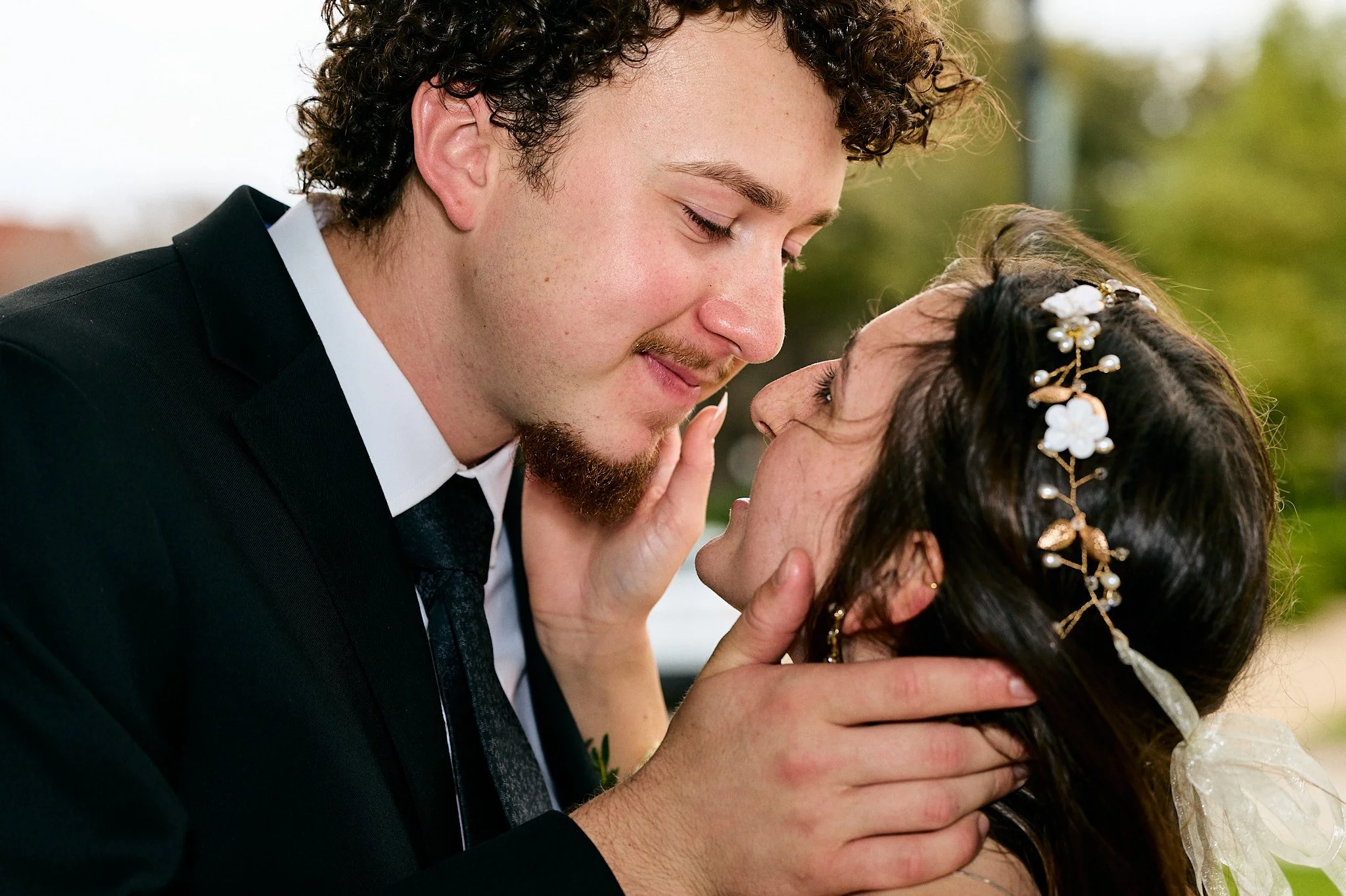 A man and a woman are sharing a romantic moment, with their foreheads touching and eyes closed. The man has curly hair, a beard, and is wearing a black suit and white shirt. The woman has dark hair adorned with a decorative hairpiece with flowers and