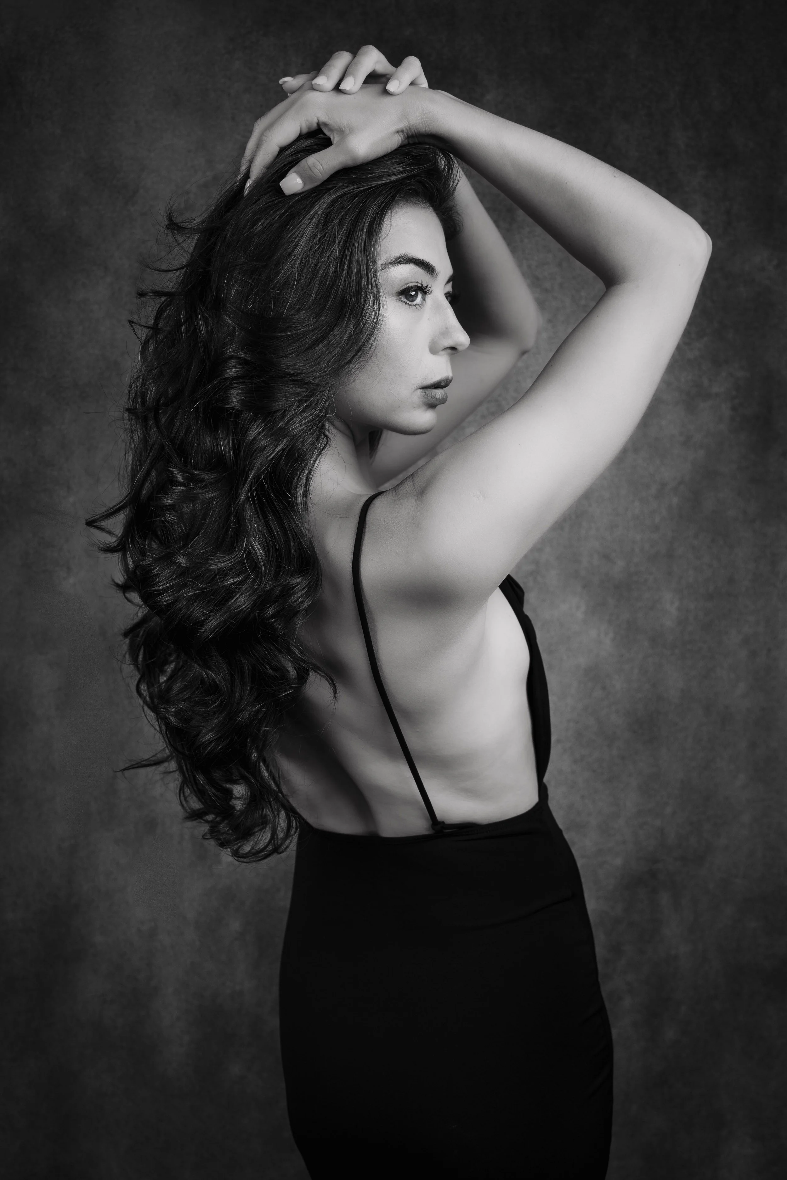 A black and white photo of a woman with long wavy hair, looking to the side, with her hands behind her head, wearing a spaghetti strap top and a high-waisted skirt.