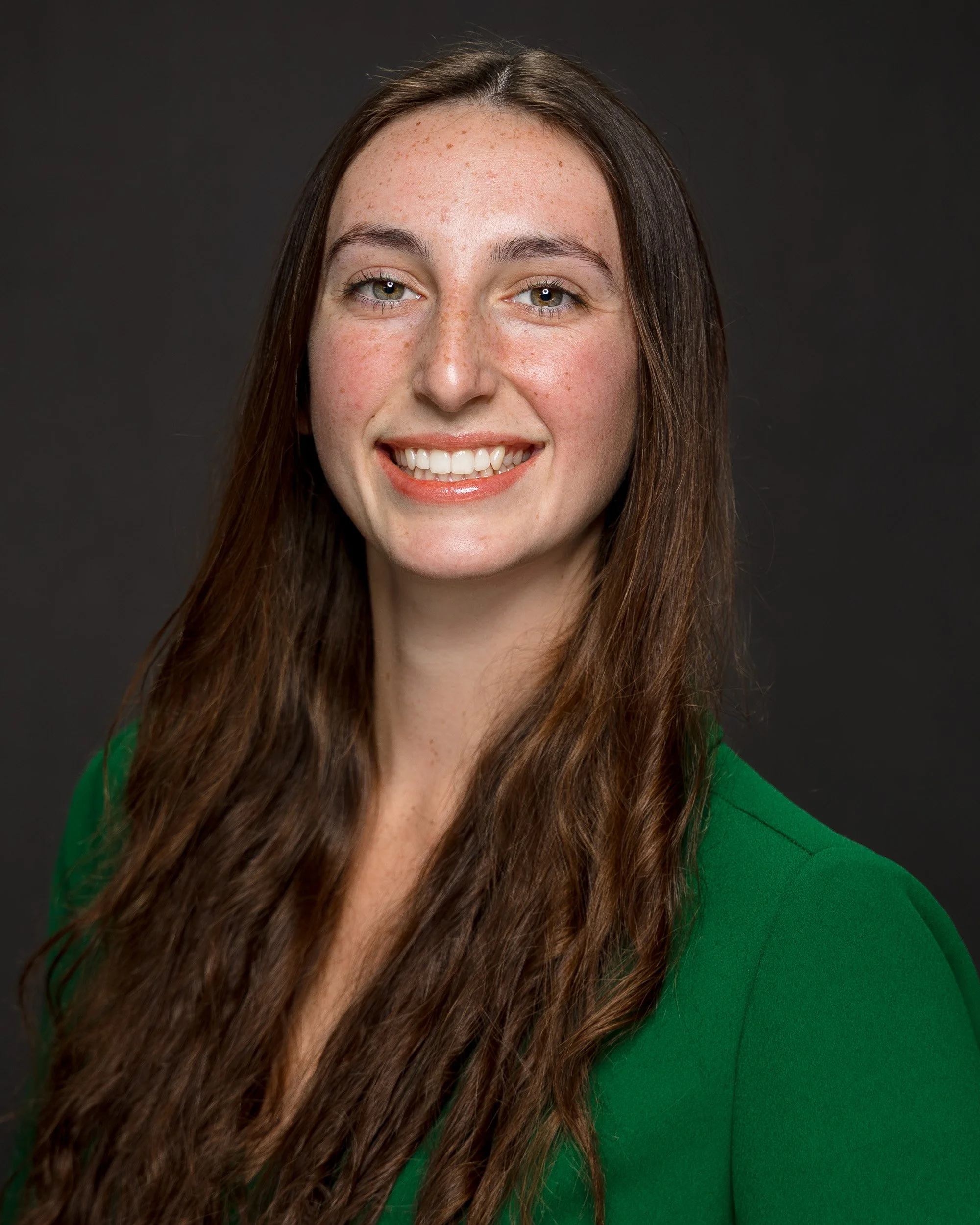 Portrait of a woman with long brown hair, light skin, freckles, wearing a green top, smiling against a dark background.