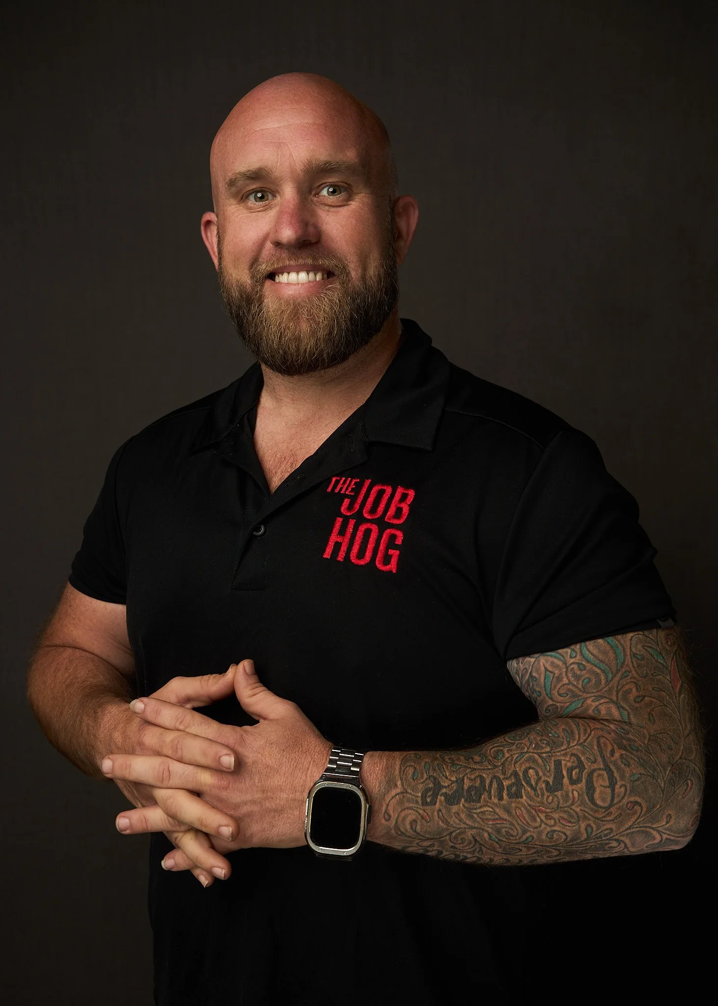 A bald man with a beard smiling, wearing a black shirt with red text, a tattoo sleeve on his right arm, a digital watch, posing against a dark background.