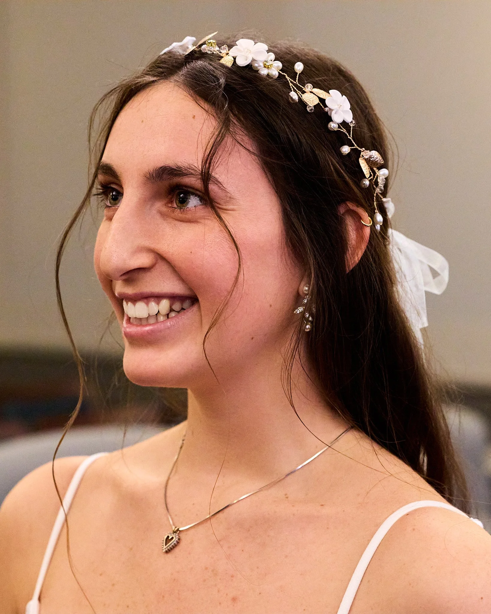 Close-up of a woman smiling with a floral headpiece, visible earrings, a heart-shaped pendant necklace, and white straps, against a neutral background.