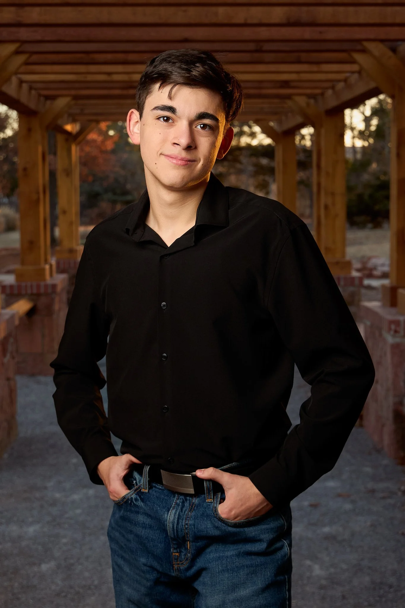 Young man wearing black shirt and blue jeans standing outdoors under a wooden archway with trees and sunset in the background.