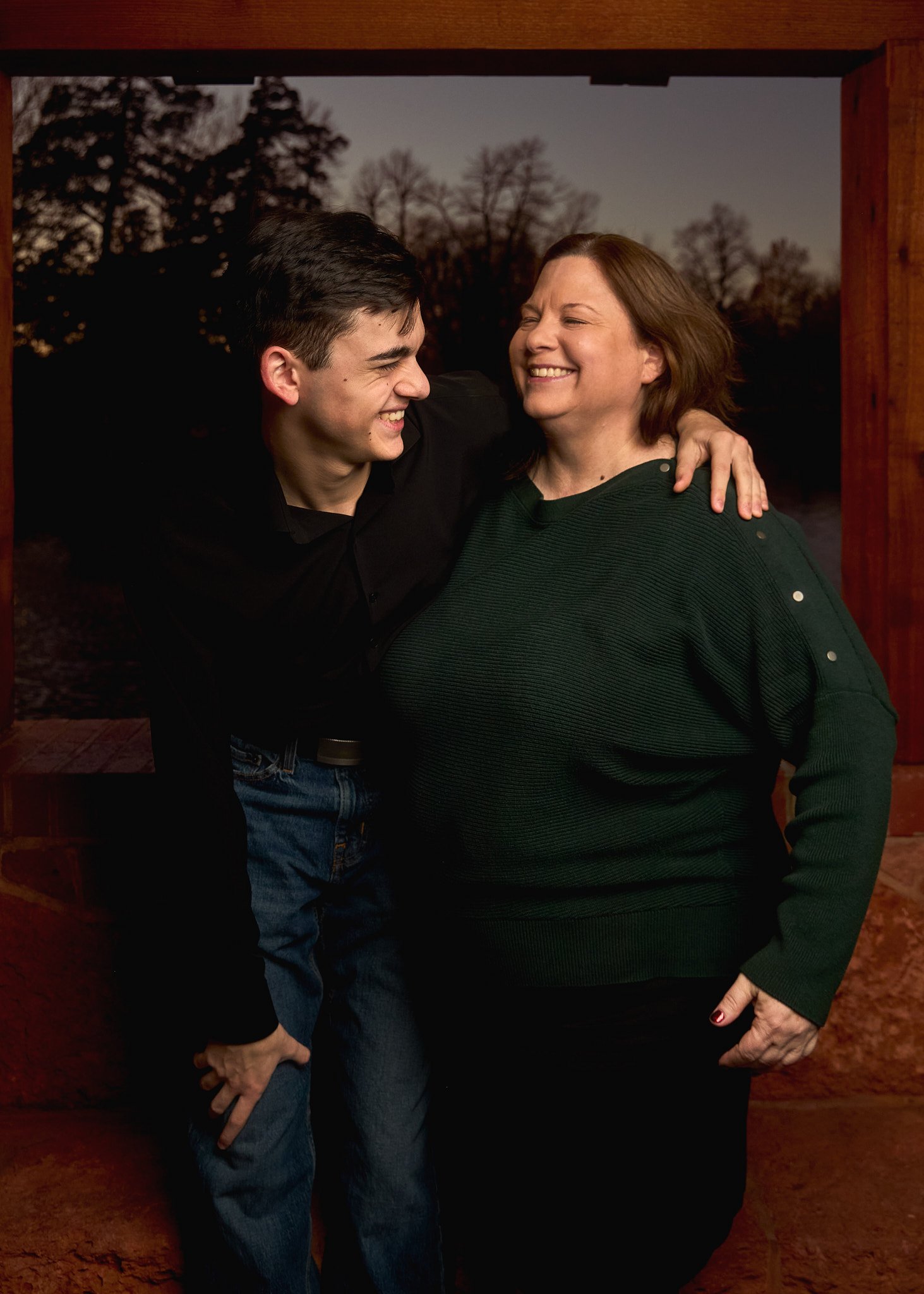 A young man and a woman standing close together outdoors at sunset, smiling and laughing, with trees in the background.