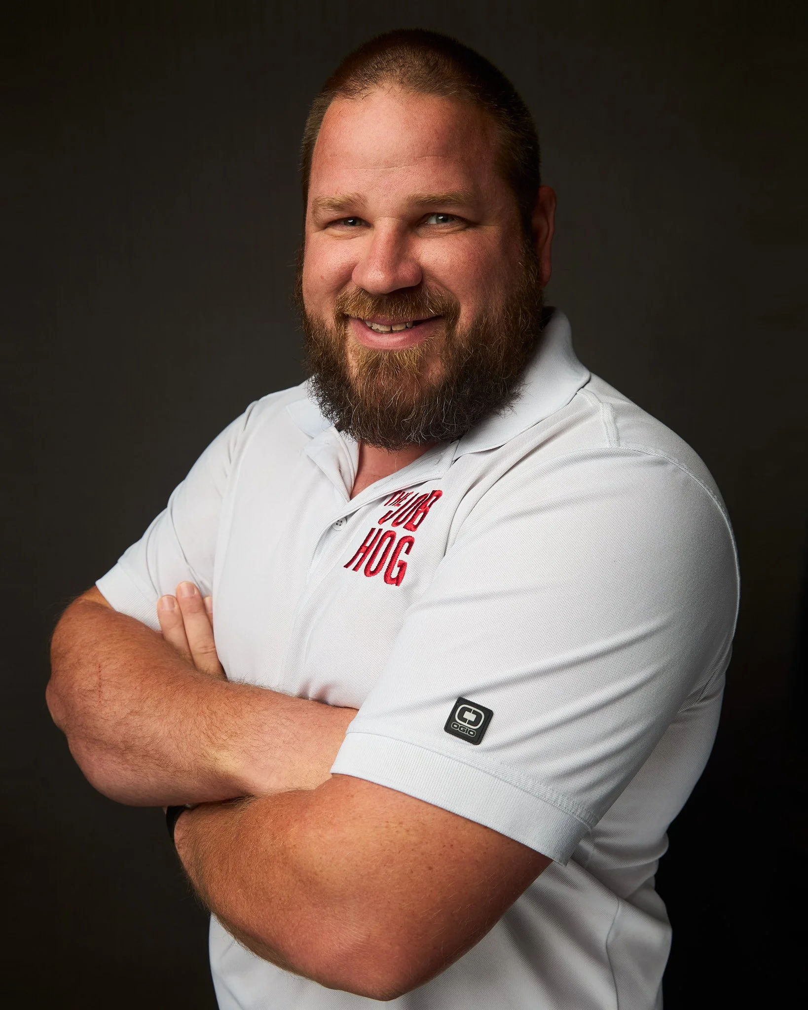 A smiling man with a beard and short hair wearing a white Polo shirt crossing his arms against a dark background.