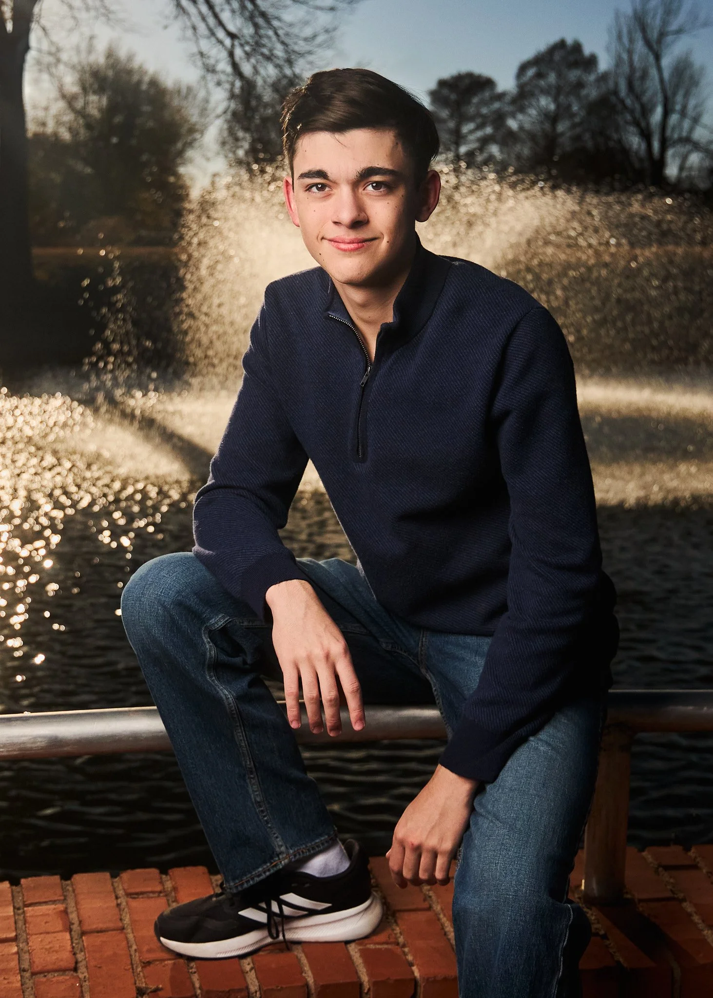 A young man with dark hair and light skin sitting outdoors on a brick surface near a body of water and trees. He is wearing a dark blue quarter-zip sweater, jeans, and black sneakers with white soles. The background features water sparkling in the su