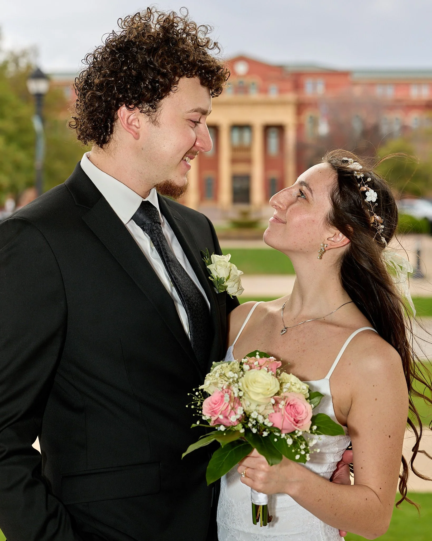 A wedding couple smiling and looking at each other outdoors, with a brick building and trees in the background. The groom wears a black suit, white shirt, and tie, and the bride wears a white dress and floral hair accessories, holding a bouquet of wh