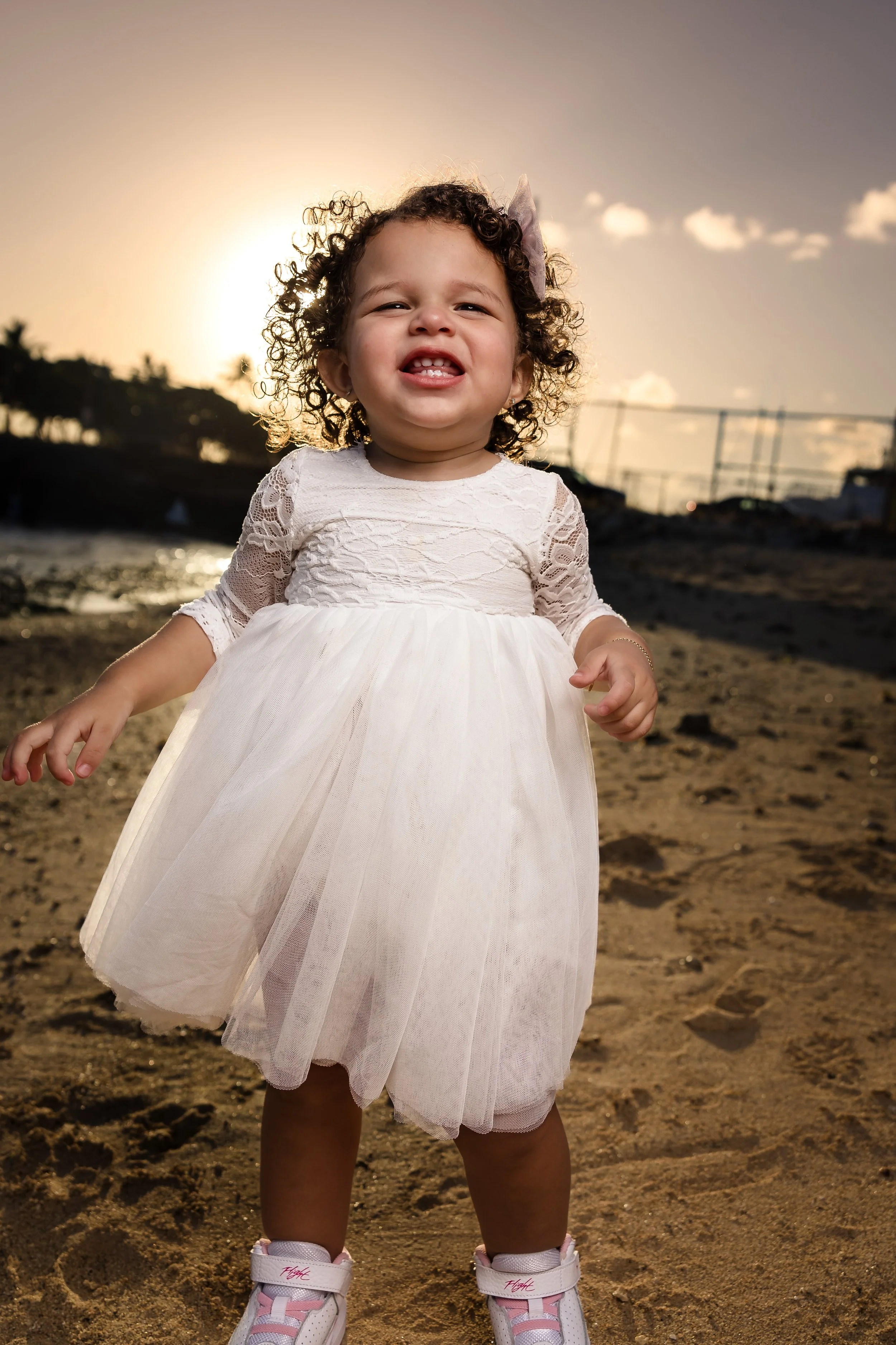 A young girl with curly hair, wearing a white lace dress and pink sneakers, standing on the beach at sunset, smiling and showing her teeth.