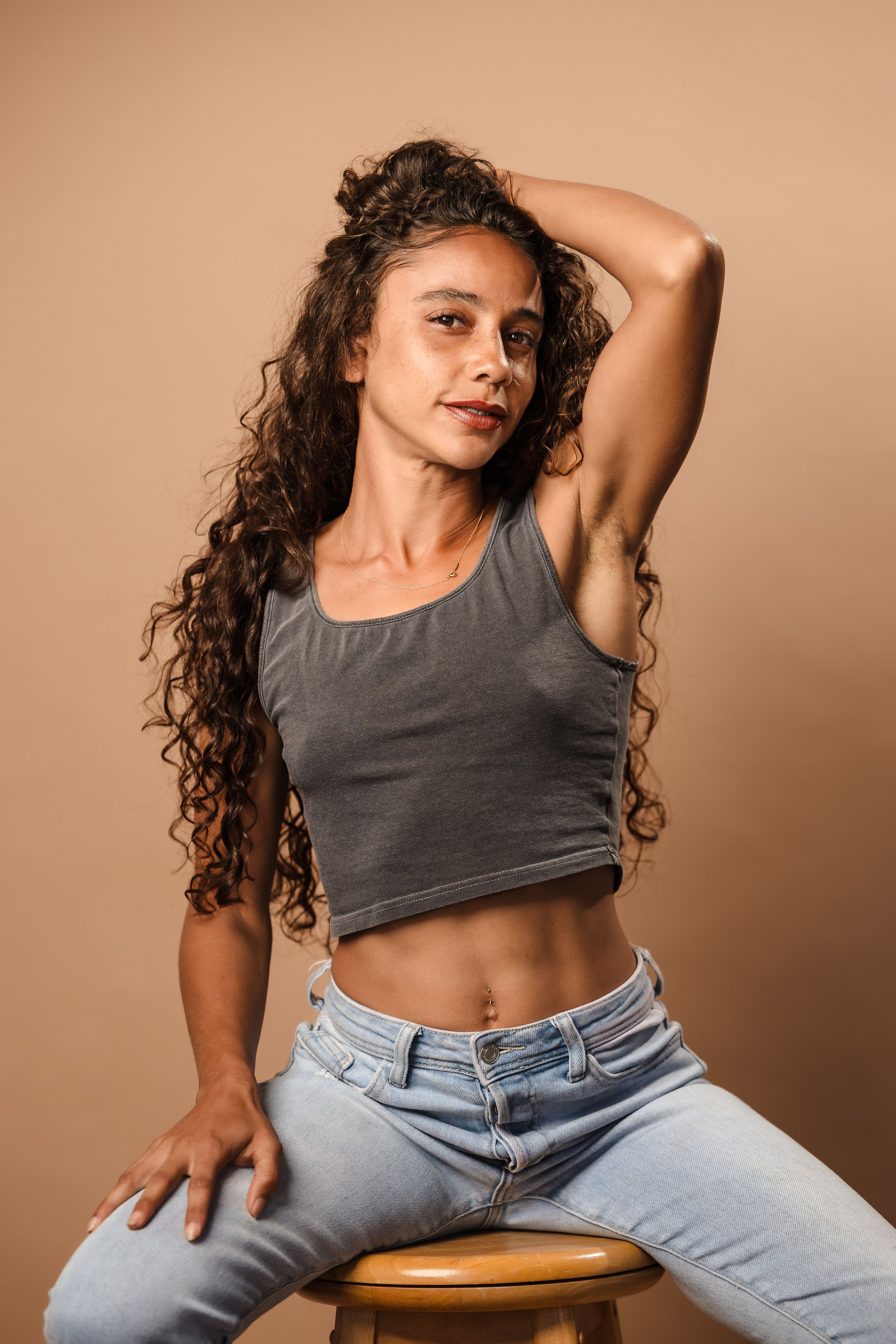 Young woman with curly hair in a gray tank top and light blue jeans, sitting on a wooden stool, with one arm raised and hand behind her head, against a beige background.