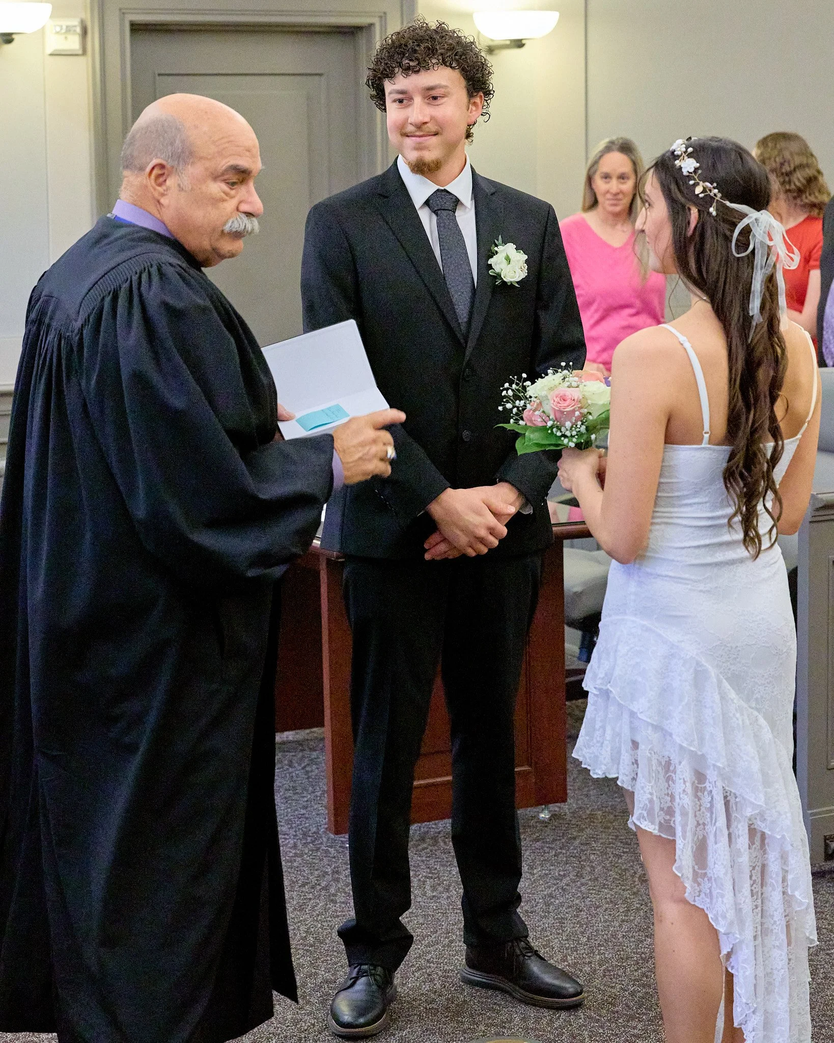 A wedding officiant, bride, and groom standing together during a wedding ceremony. The officiant is reading from a small booklet, the groom is dressed in a black suit with a boutonniere, and the bride in a white lace dress with floral hair accessorie