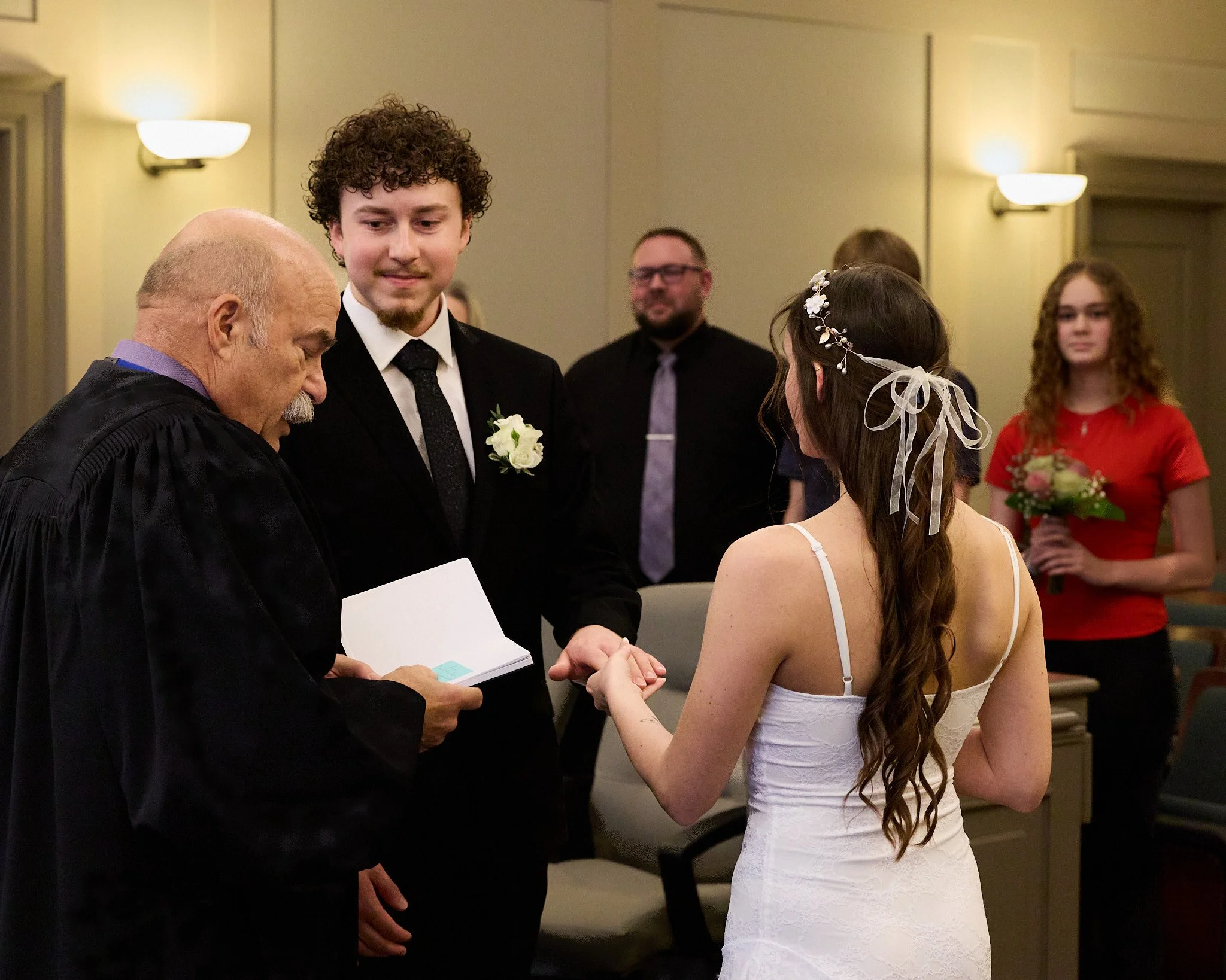 A young woman with long, wavy brown hair wearing a white dress and floral hair accessory during a wedding ceremony, exchanging rings with a man in a black suit, with several people watching in the background.