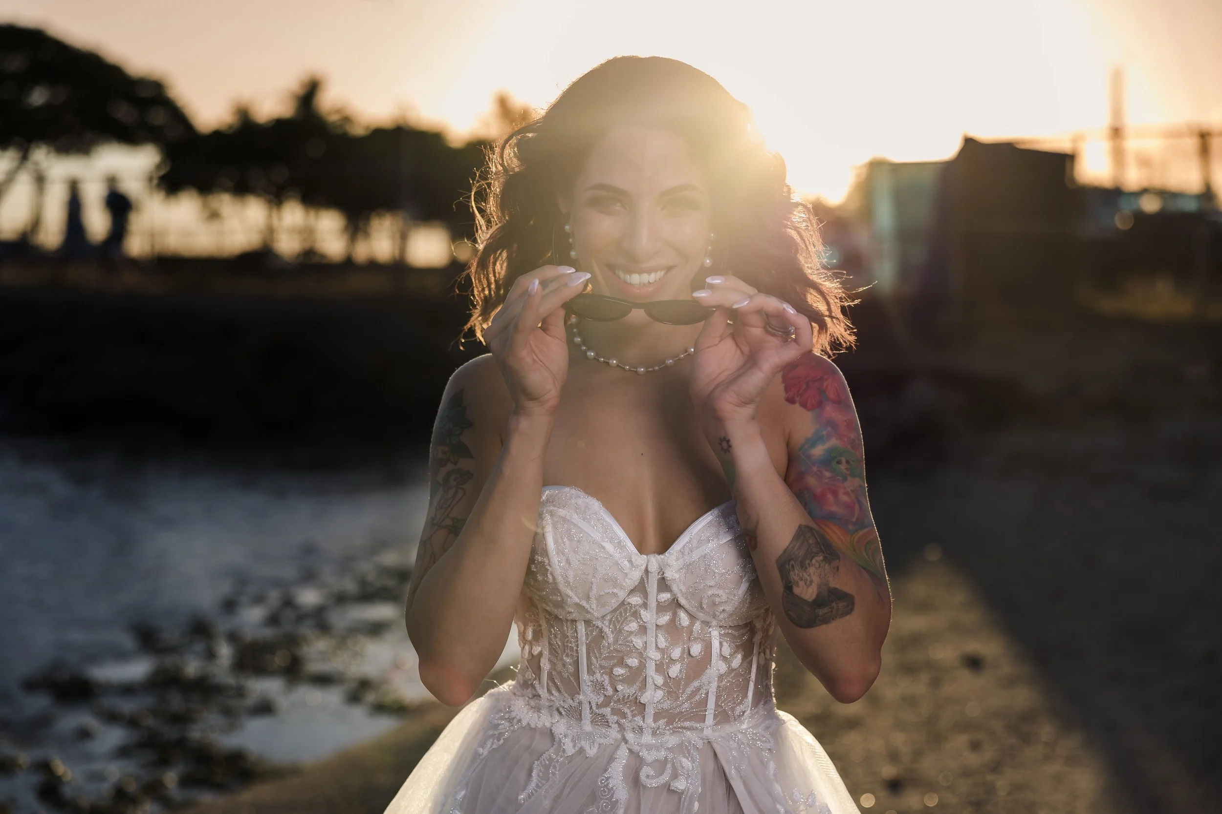 A woman in a wedding dress holding sunglasses, smiling at the camera, with tattoos on her arms, during sunset on a beach.