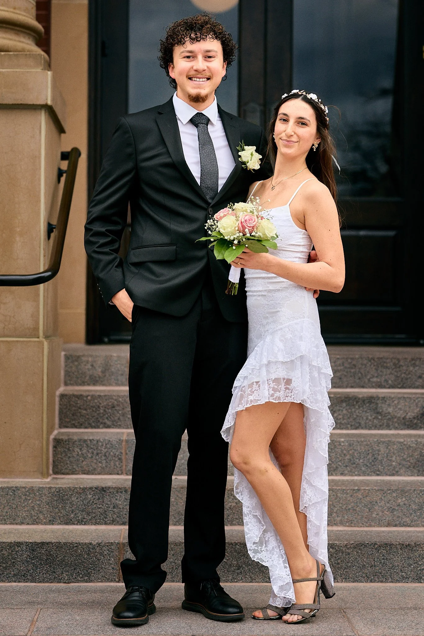 A smiling man in a black suit and a woman in a white lace dress holding a bouquet of pink and white flowers, standing on the steps outside a building.
