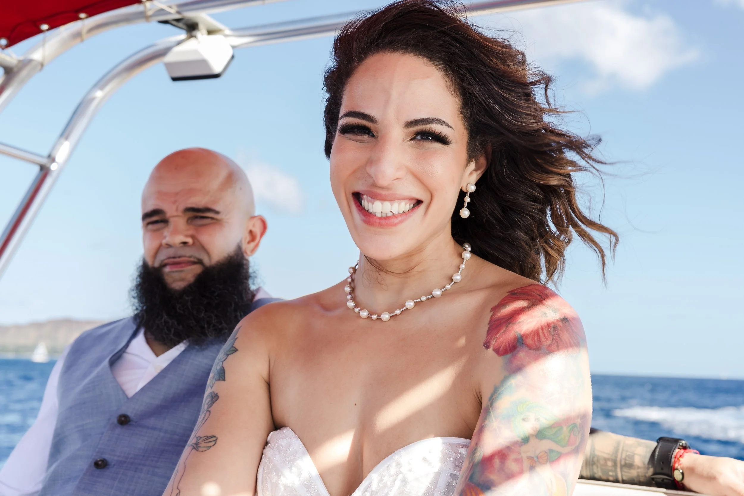 A smiling woman with dark hair, tattoos, and pearl jewelry, wearing a strapless dress, is in a boat with a man with a beard and bald head, dressed in a gray suit, against a bright blue sky and water background.