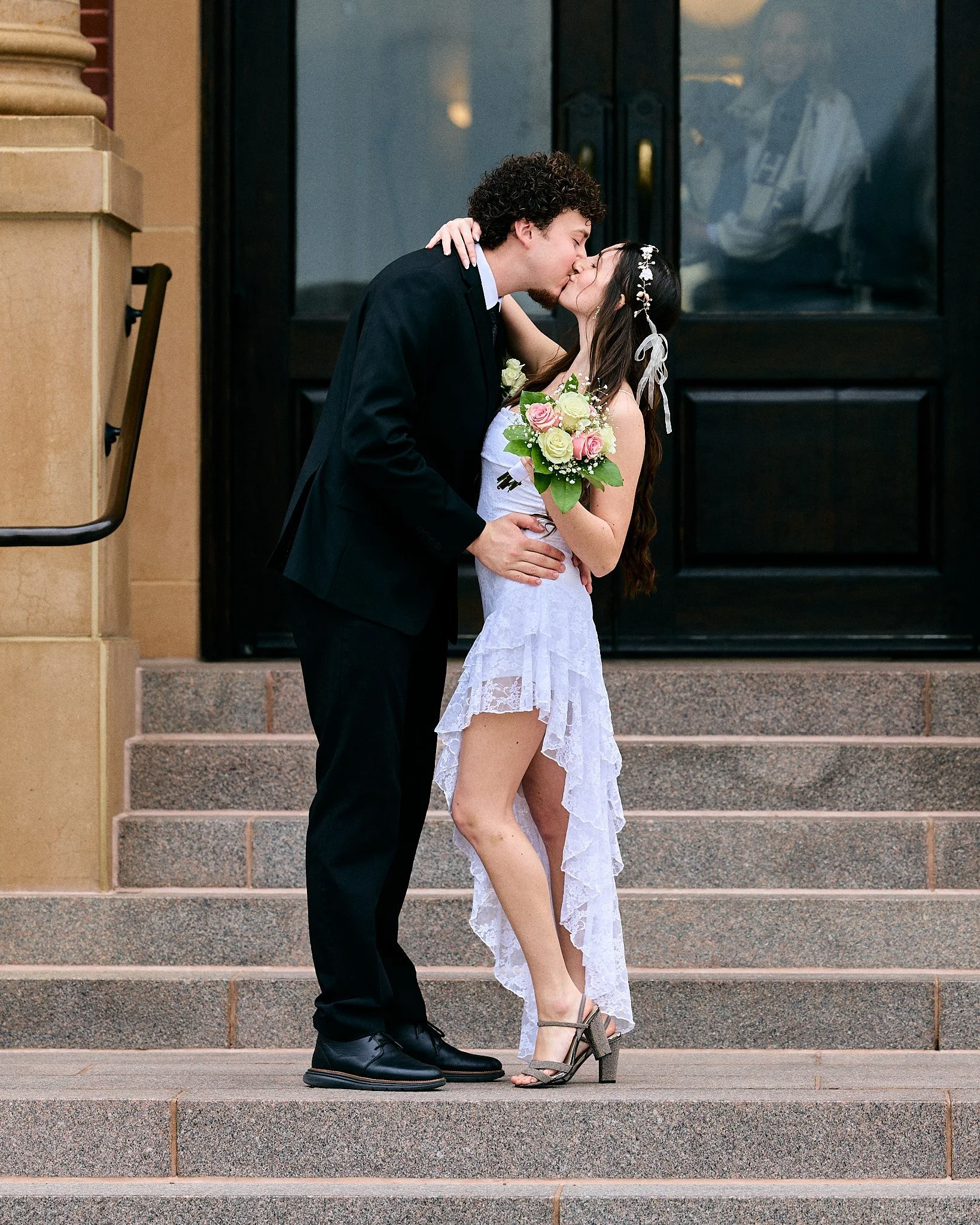 A couple kissing on the steps outside of a building, with the woman holding a bouquet of flowers and wearing a lace dress and the man in a black suit and tie.