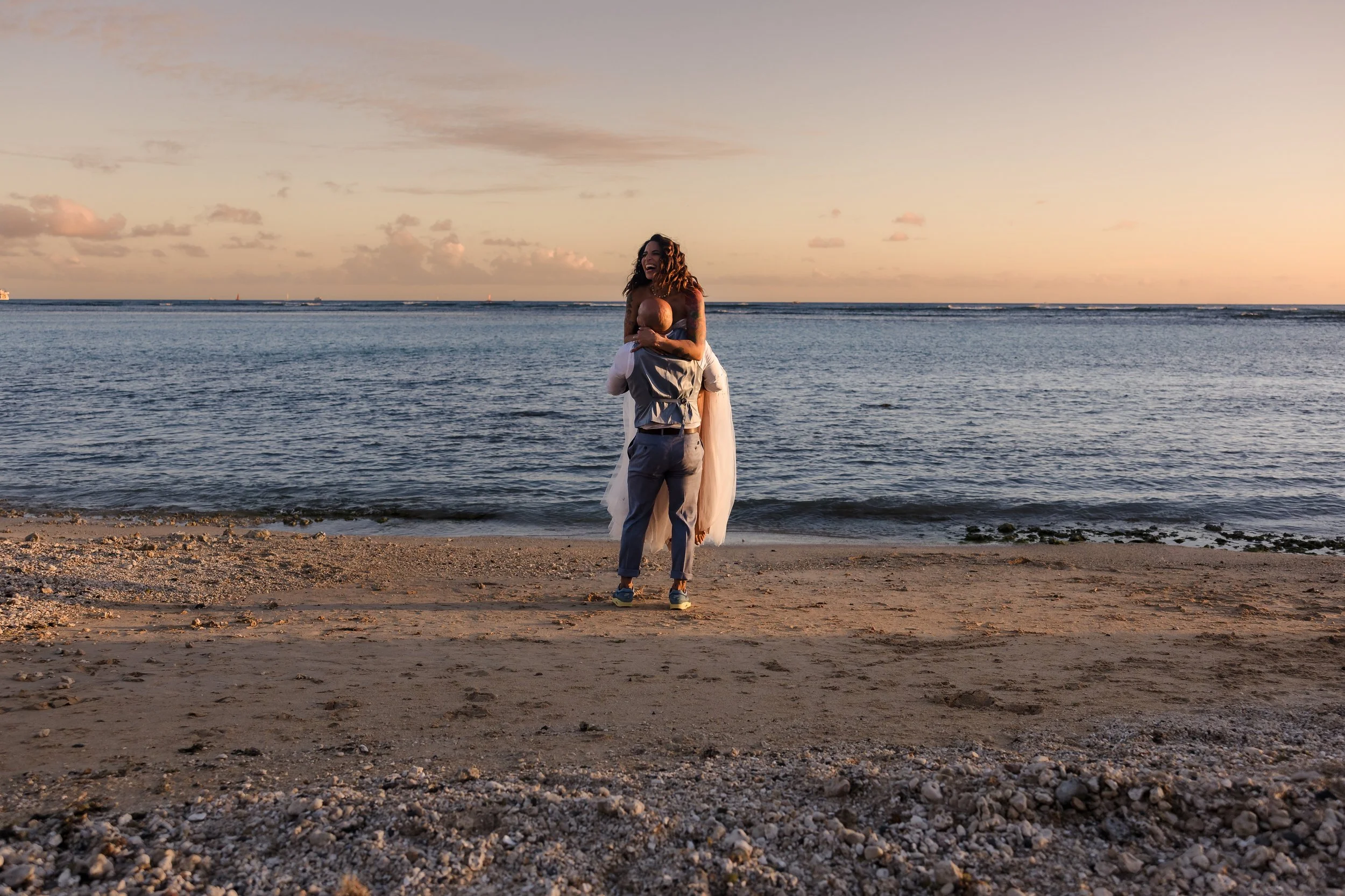A couple on the beach at sunset, with the woman sitting on the man's shoulders, both smiling and enjoying the moment.