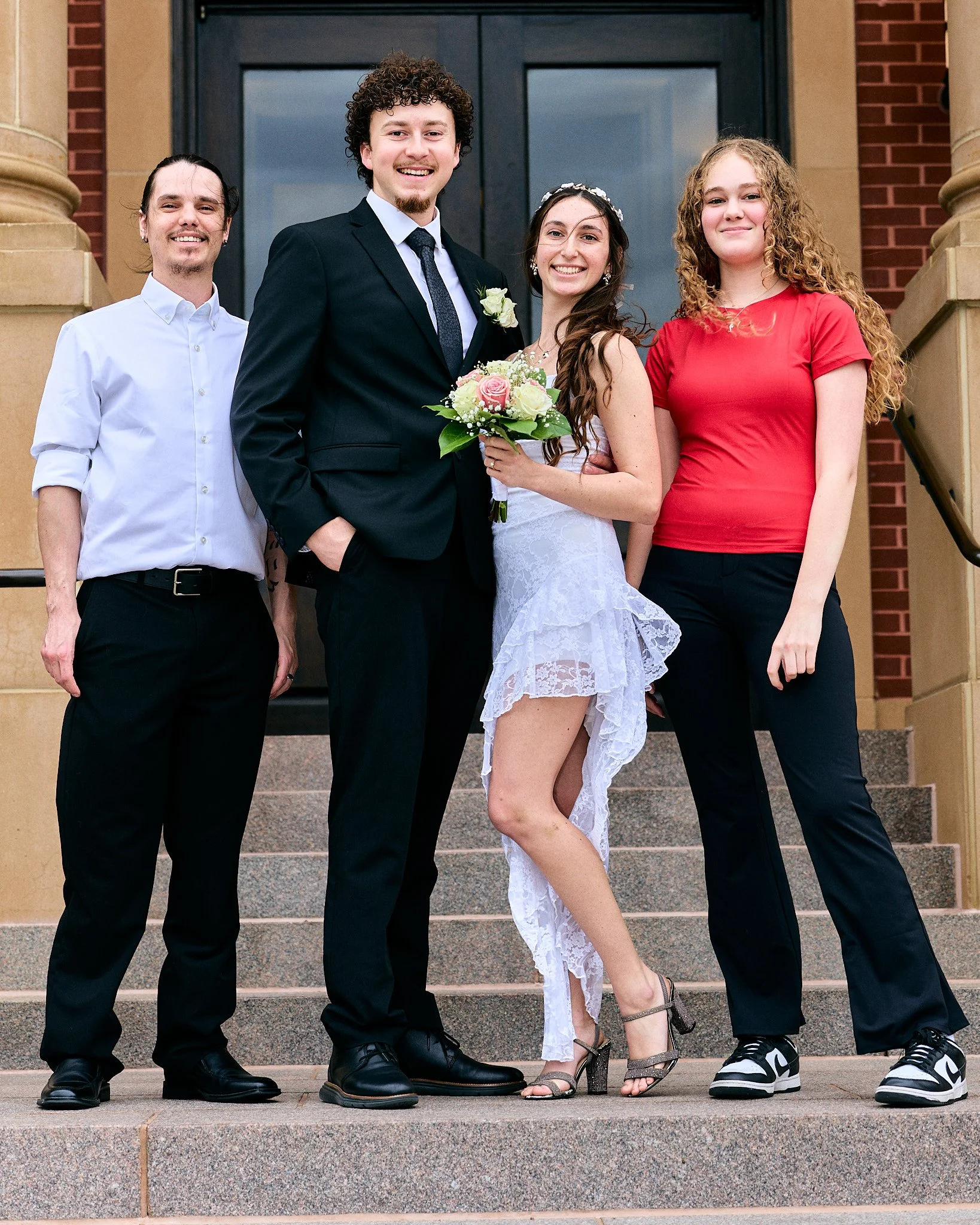 Group of five young adults standing on steps outside building, dressed for a wedding, smiling at camera, one holding a bouquet of flowers.