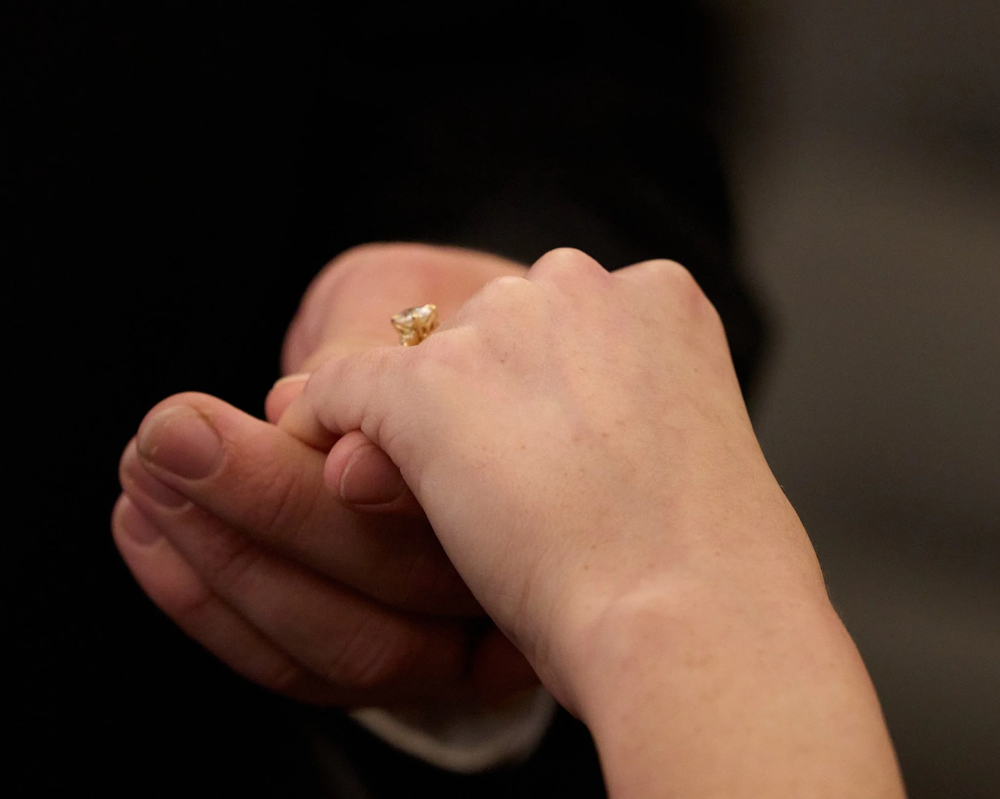 Close-up of a hand with a diamond ring on the ring finger, held and gently grasped by another person's hand against a dark background.