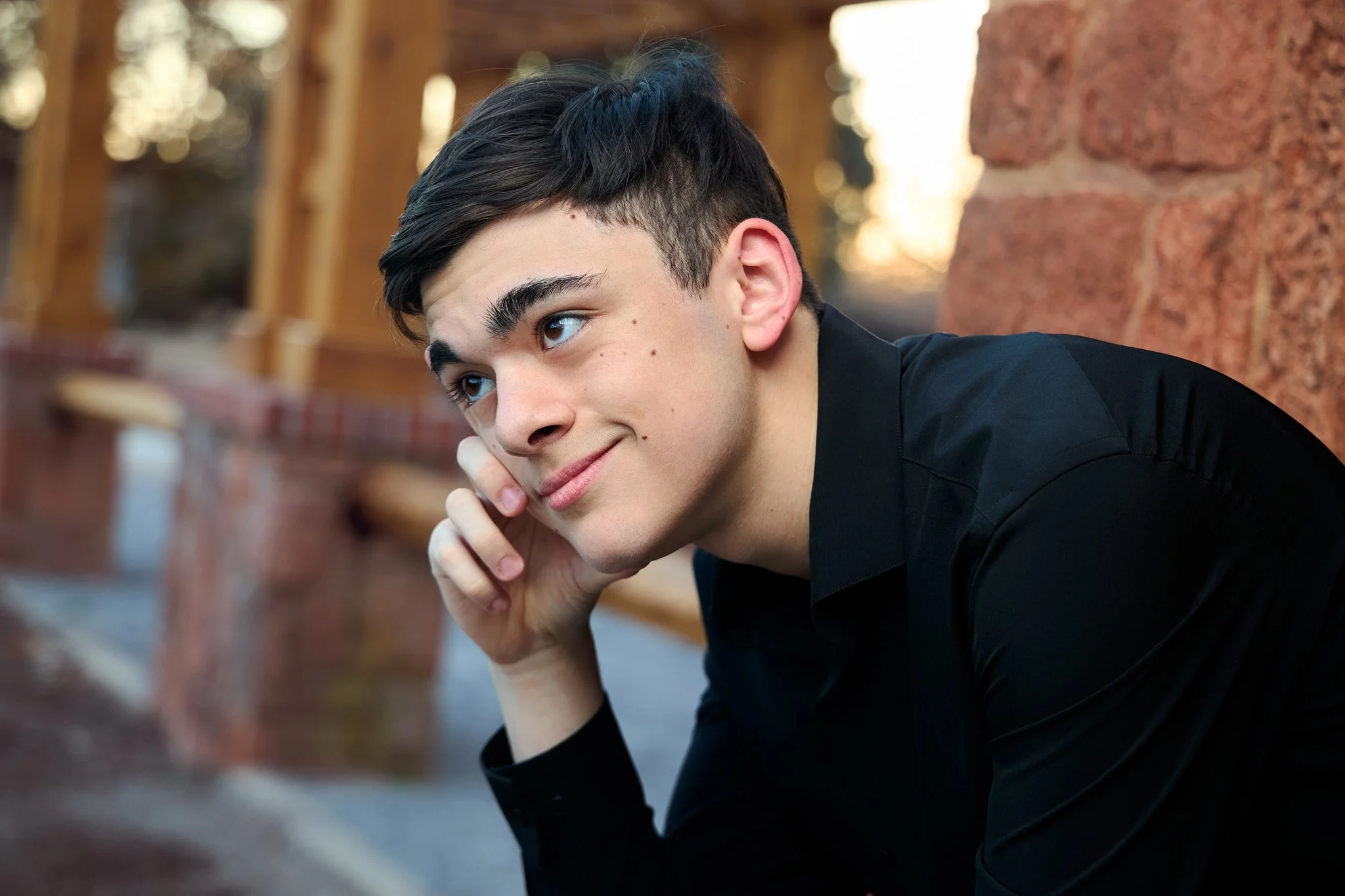 A young man with styled dark hair and a slight smile, resting his chin on his hand while looking to the side, outdoors with a blurred background of trees and a brick wall.