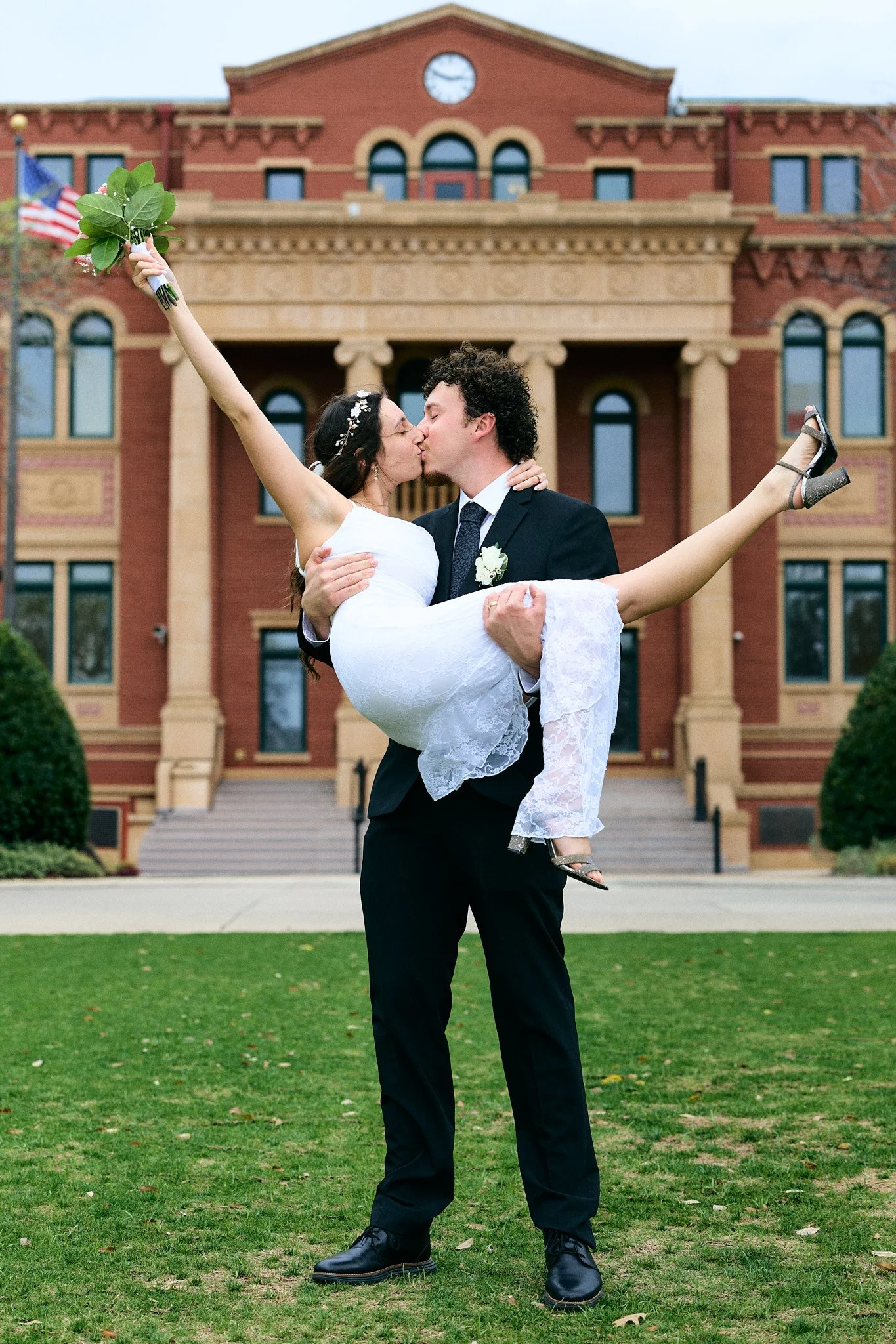 A man in a dark suit holding a woman in a white dress and silver heels in front of a large red brick building. The woman is holding a bouquet of greenery and flowers. They are kissing, with the woman extending her arm and leg outward.