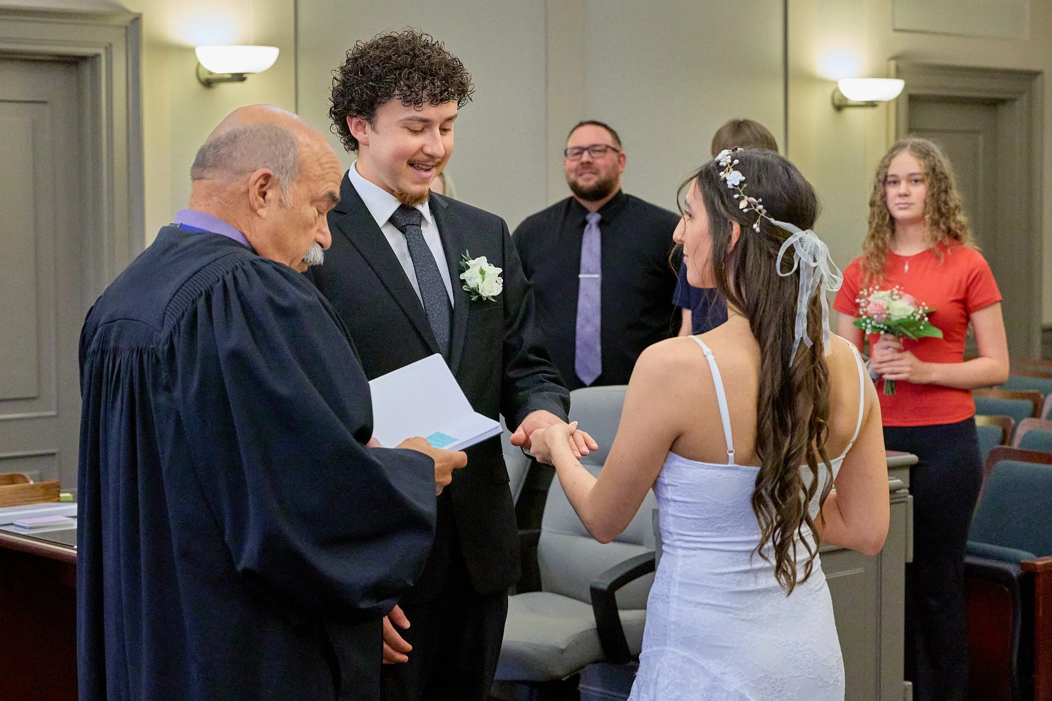 A wedding ceremony where a bride and groom are exchanging vows and rings in front of a judge or officiant, with wedding guests watching.