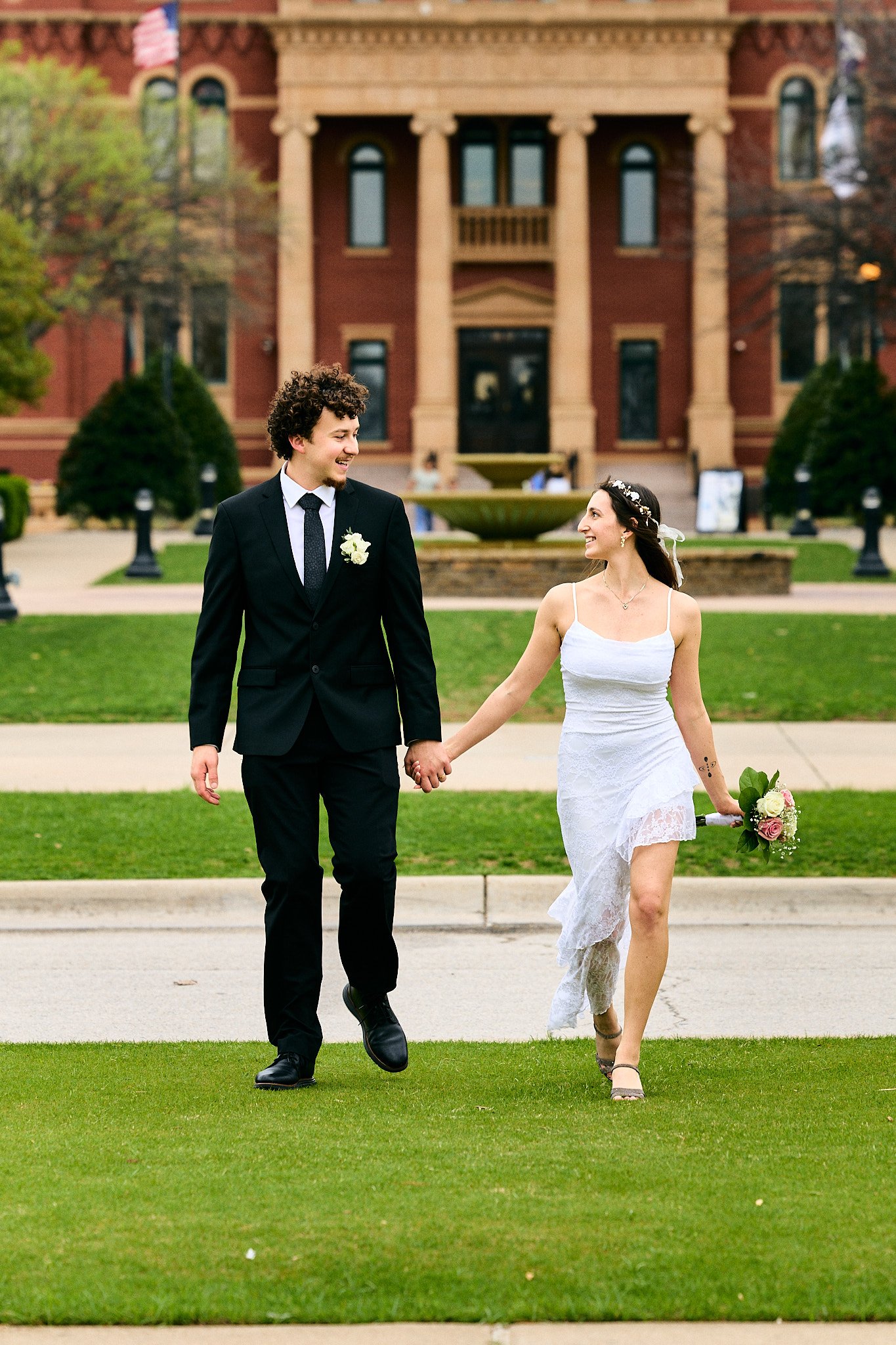 A wedding couple holding hands and walking outside in front of a large brick building, with the groom in a black suit and tie and the bride in a white lace dress holding a bouquet of flowers.