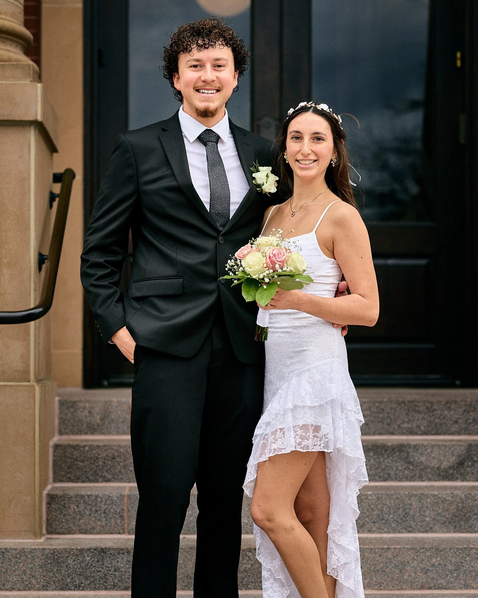 A smiling man in a black tuxedo and a woman in a white lace dress holding a bouquet of flowers, standing on steps in front of a black door.