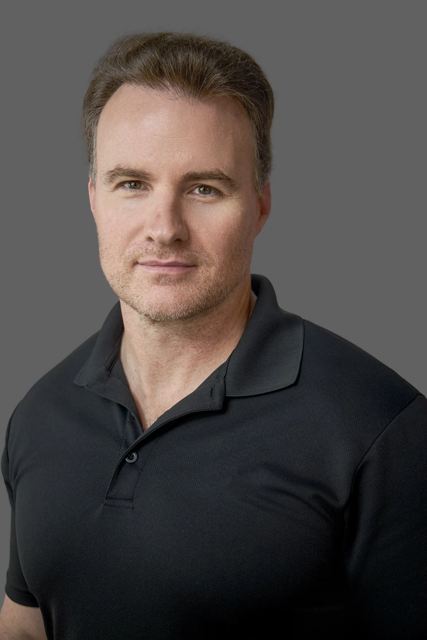 A man with short brown hair, slight facial hair, and light skin wearing a black collared polo shirt, against a plain gray background.