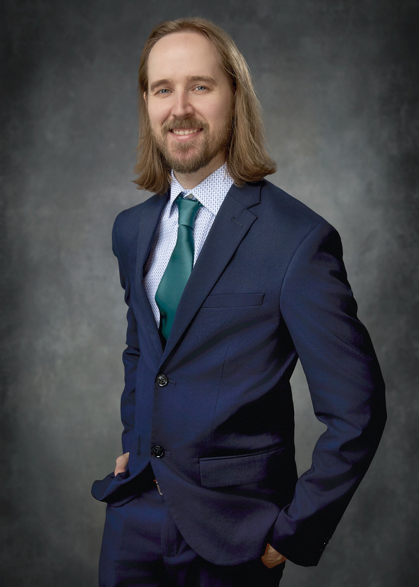 A man with shoulder-length brown hair, a beard, wearing a dark blue suit, a patterned white shirt, and a teal tie posing against a dark, textured background.