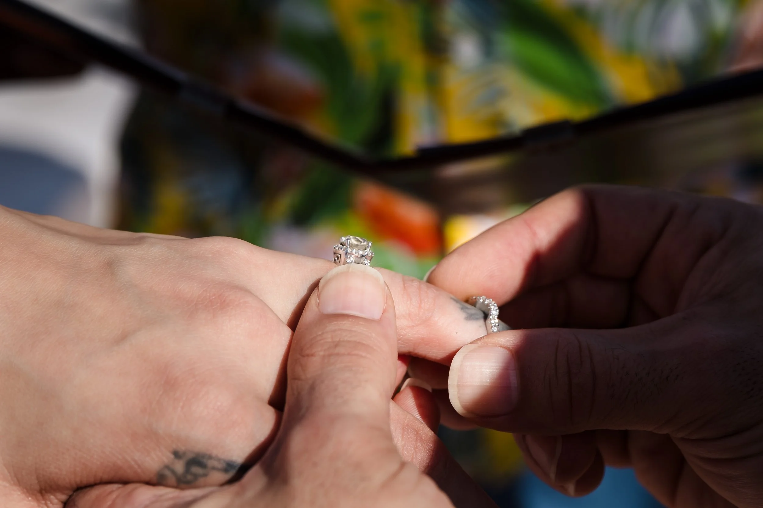 Close-up of two hands exchanging rings, one with a diamond ring, suggestive of a wedding or engagement ceremony, with a blurred colorful background.