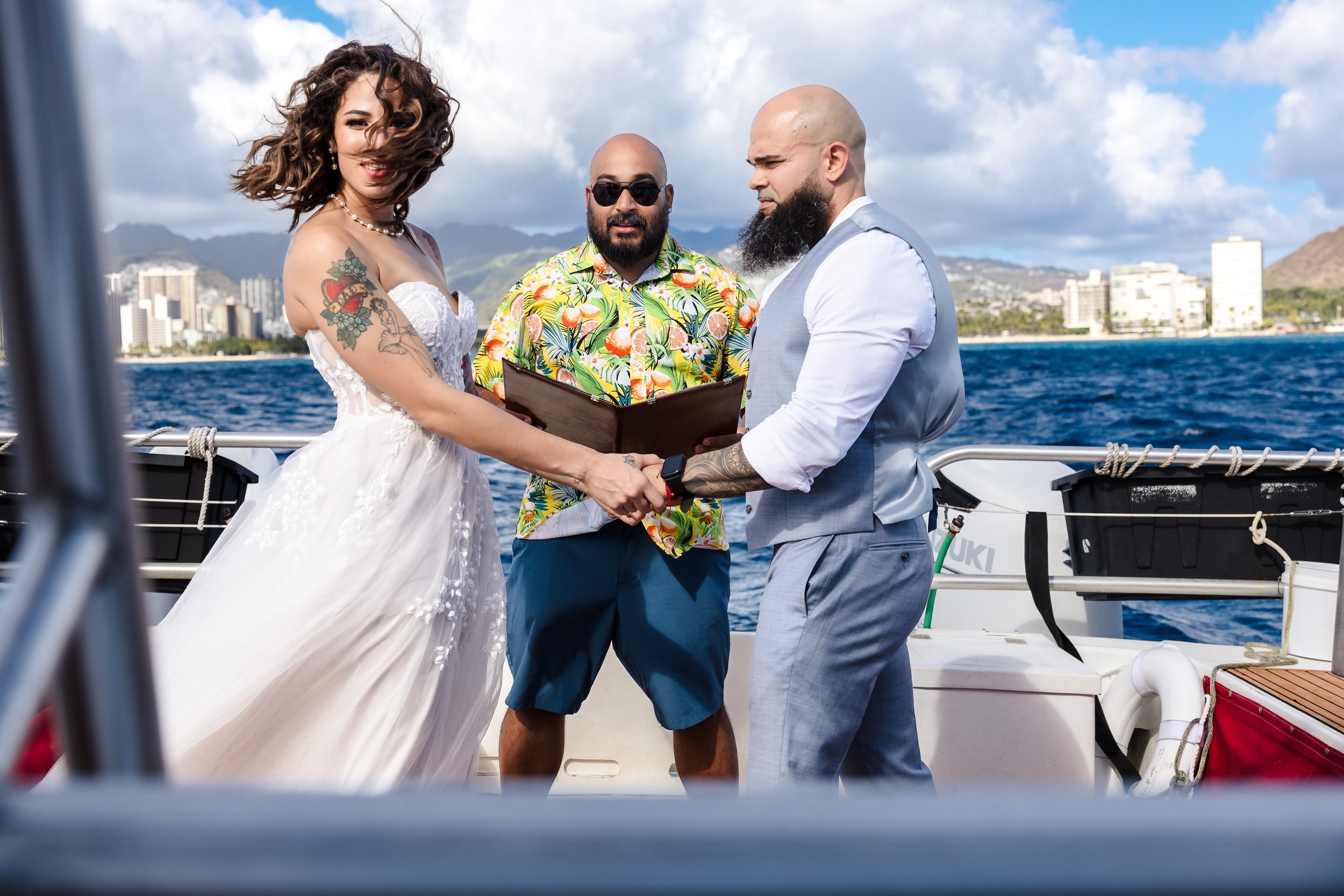 A couple getting married on a boat, holding hands with an officiant dressed in a tropical shirt between them. The bride, with curly hair, tattoos, and wearing a white dress, smiles while the groom, with a bald head, beard, and wearing a gray vest, lo