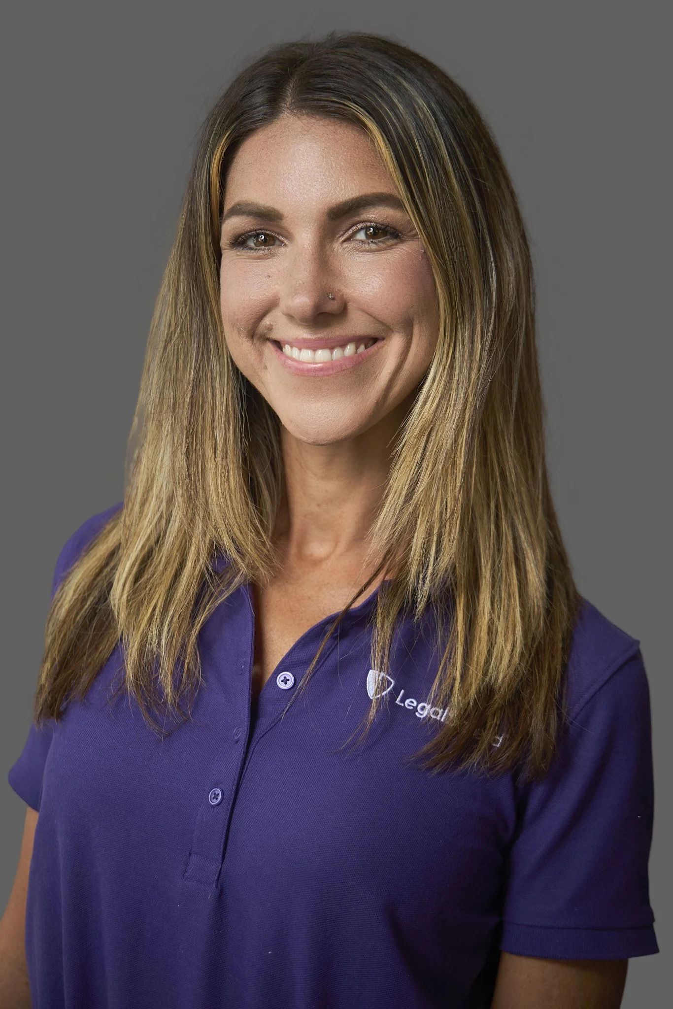 Close-up of a woman smiling, wearing a purple shirt with a logo and text partially visible on the chest, with long brown hair with lighter highlights, against a plain gray background.