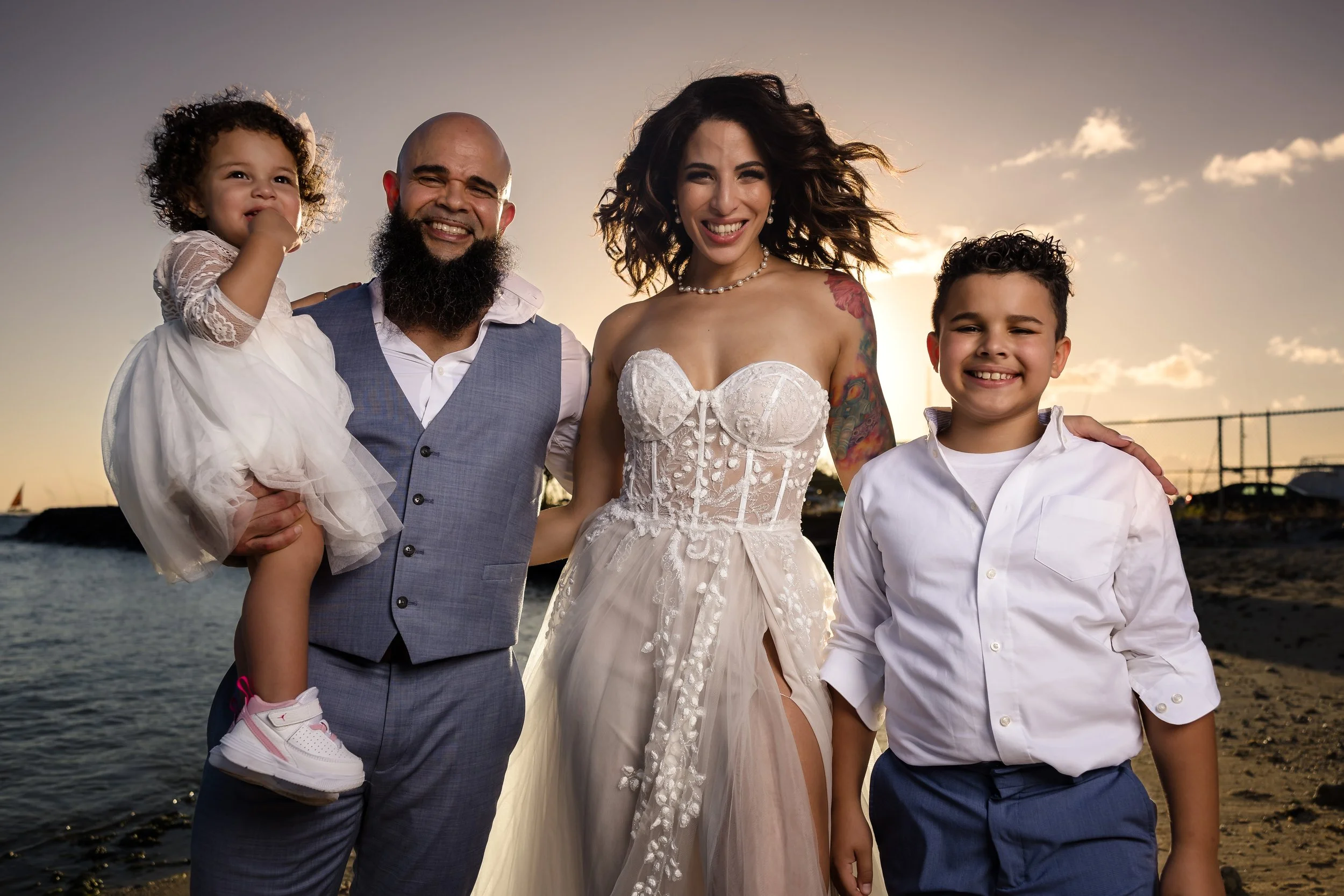 A group of five people, including a man, a woman in a wedding dress, and three children, standing on a beach during sunset, smiling.