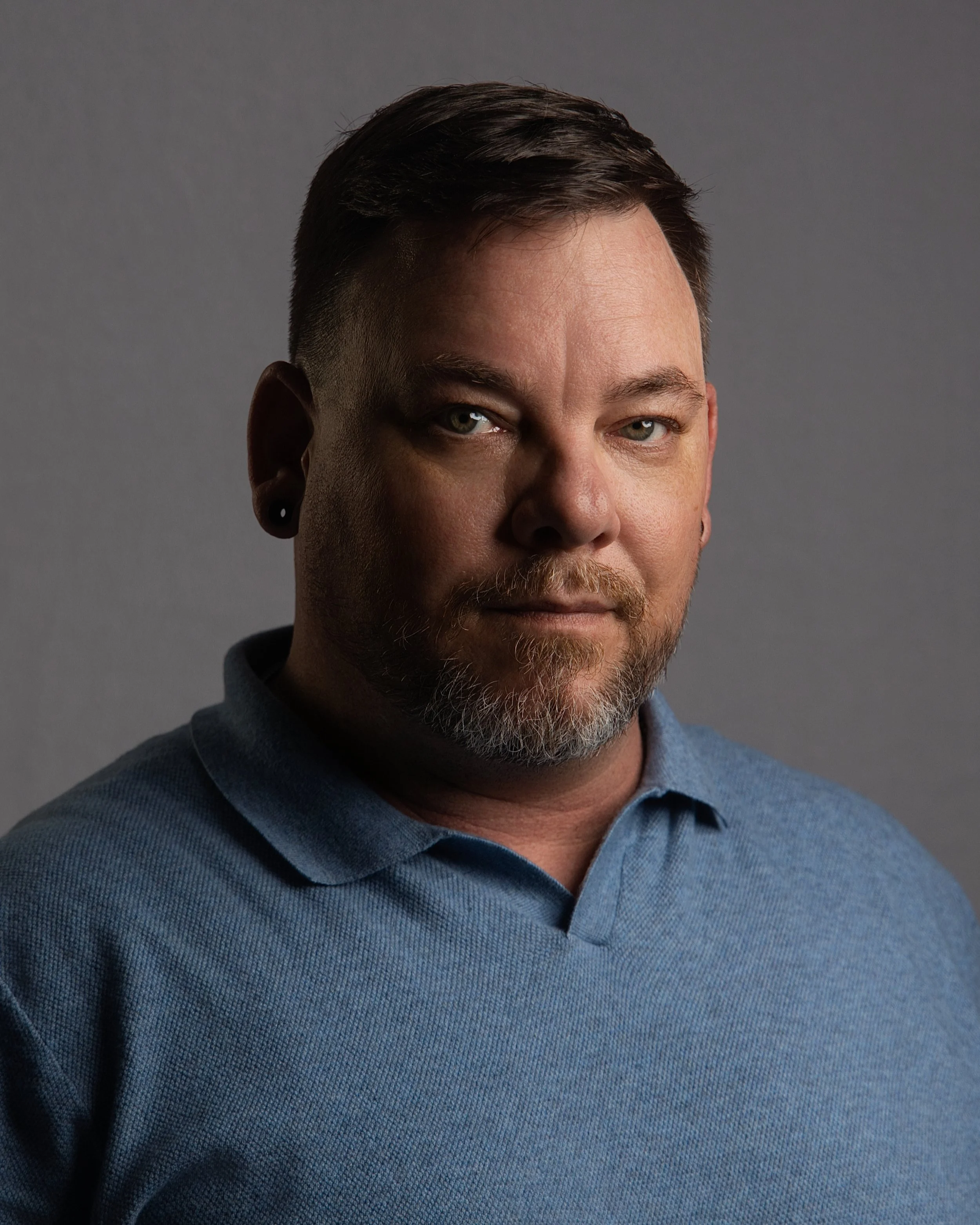 A close-up portrait of a man with light skin, blue eyes, short dark hair, a beard, and wearing a blue collared shirt, against a gray background.