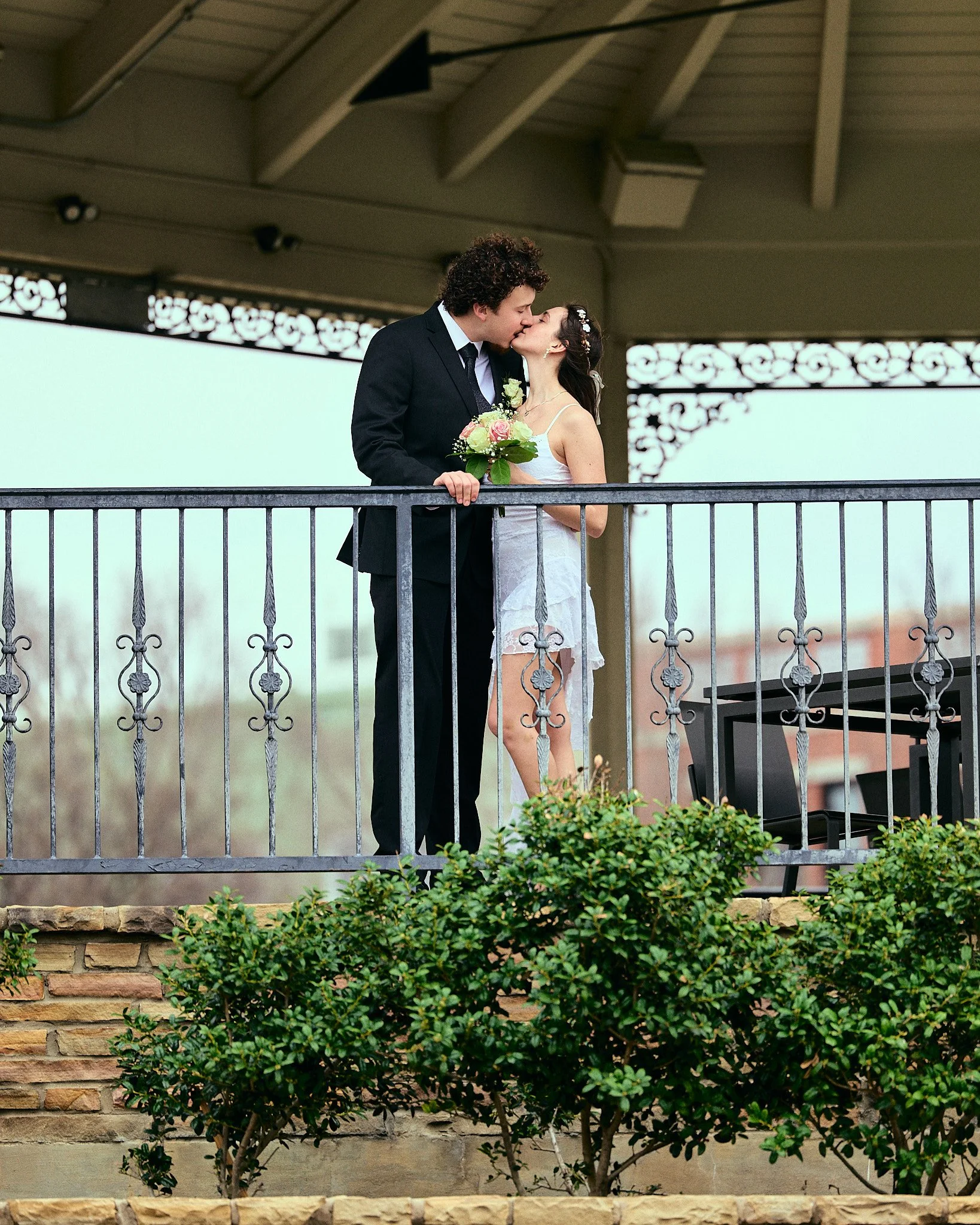 A bride and groom kissing on a balcony, with the groom in a black suit and the bride in a white dress holding a bouquet, under a covered structure with decorative ironwork.