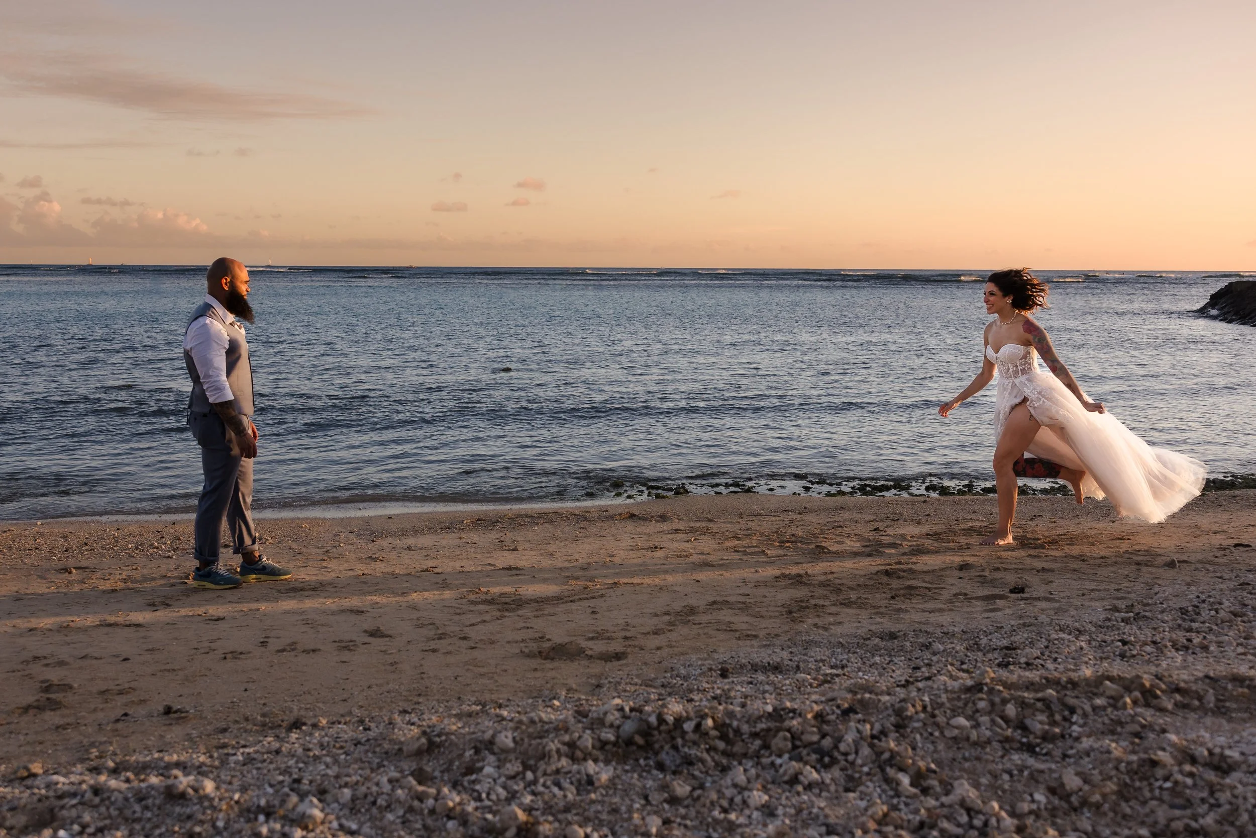 A man with a beard and tattoos on his arms stands on a beach, holding hands with a woman in a white wedding dress who is smiling and walking barefoot on the sand during sunset.