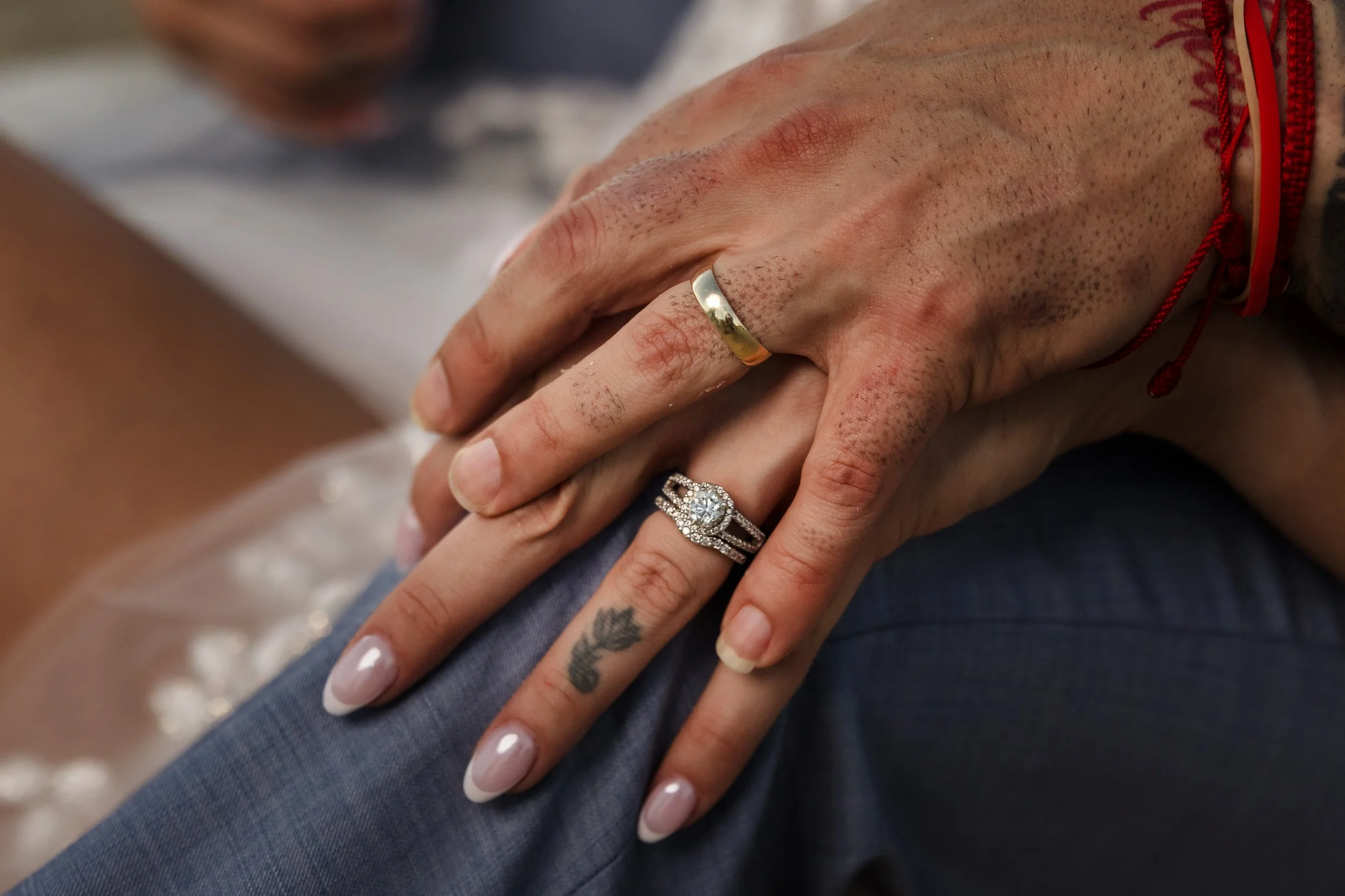 Close-up of two people's hands, one with a wedding ring, resting on a blue fabric. One hand has a tattoo and there are red string bracelets on one wrist.