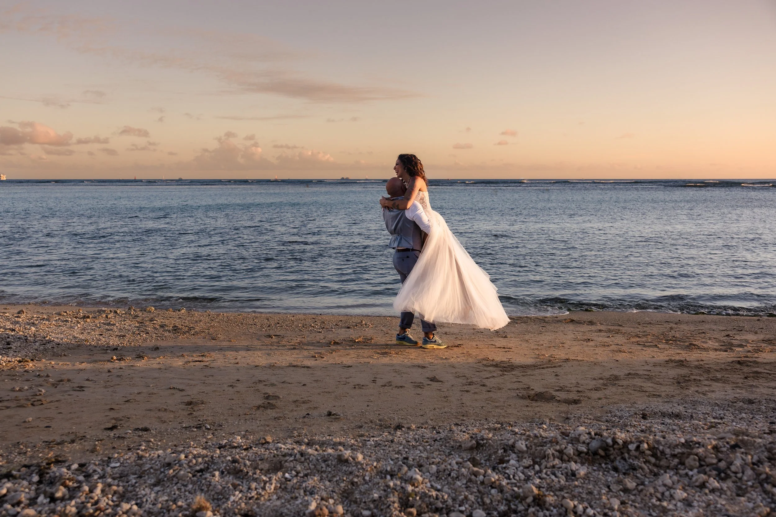 A couple is embracing on the beach at sunset, with the woman wearing a white wedding dress and the man in casual attire. The ocean and sky are visible in the background.