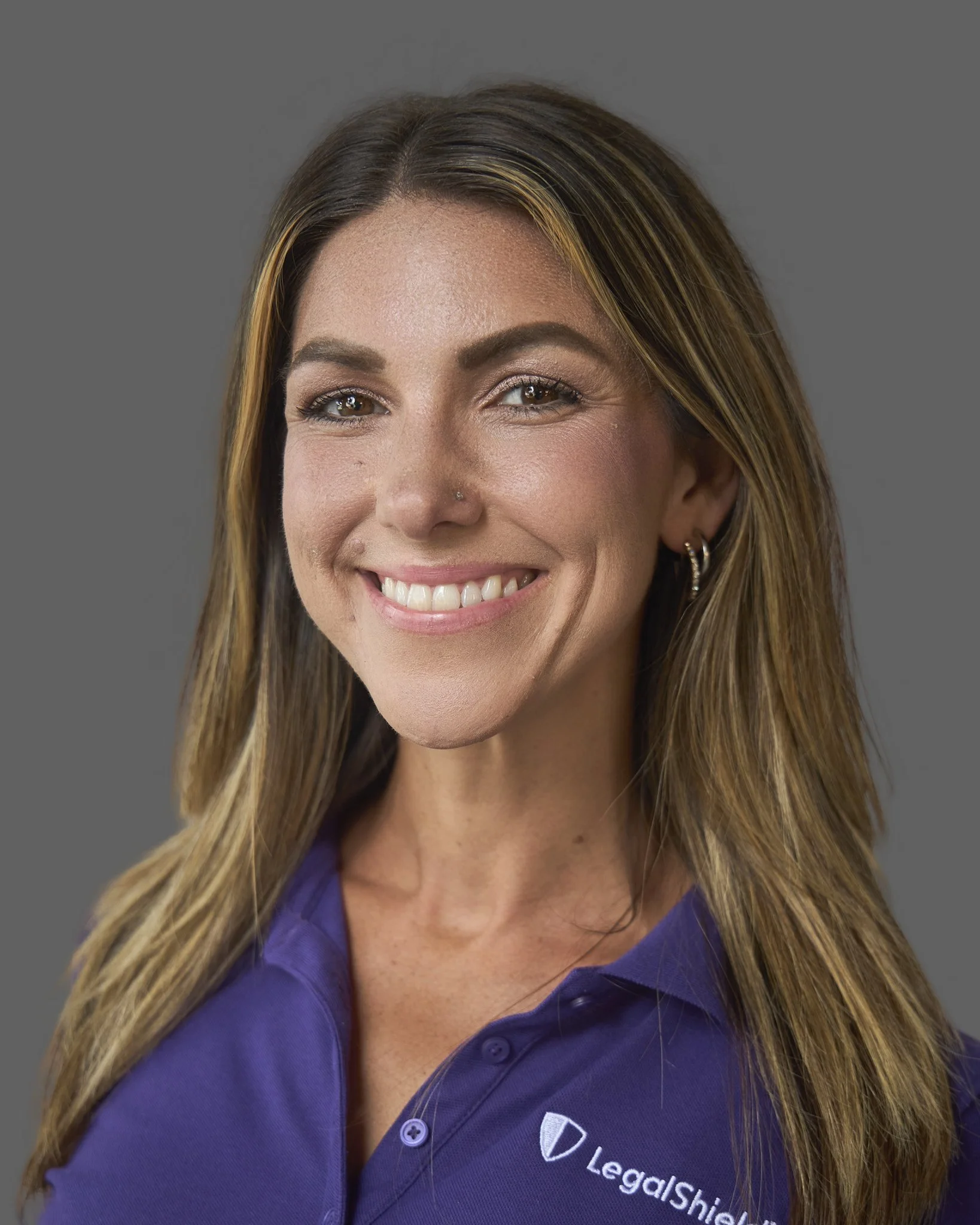 Close-up of a smiling woman with long brown hair and brown eyes, wearing a purple polo shirt with a logo and text reading 'LegalShield' on it.