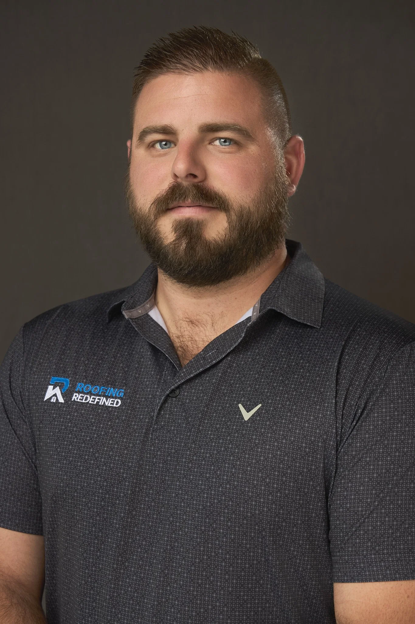 Headshot of a man with a beard and mustache, wearing a black collared shirt with logos, against a dark background.