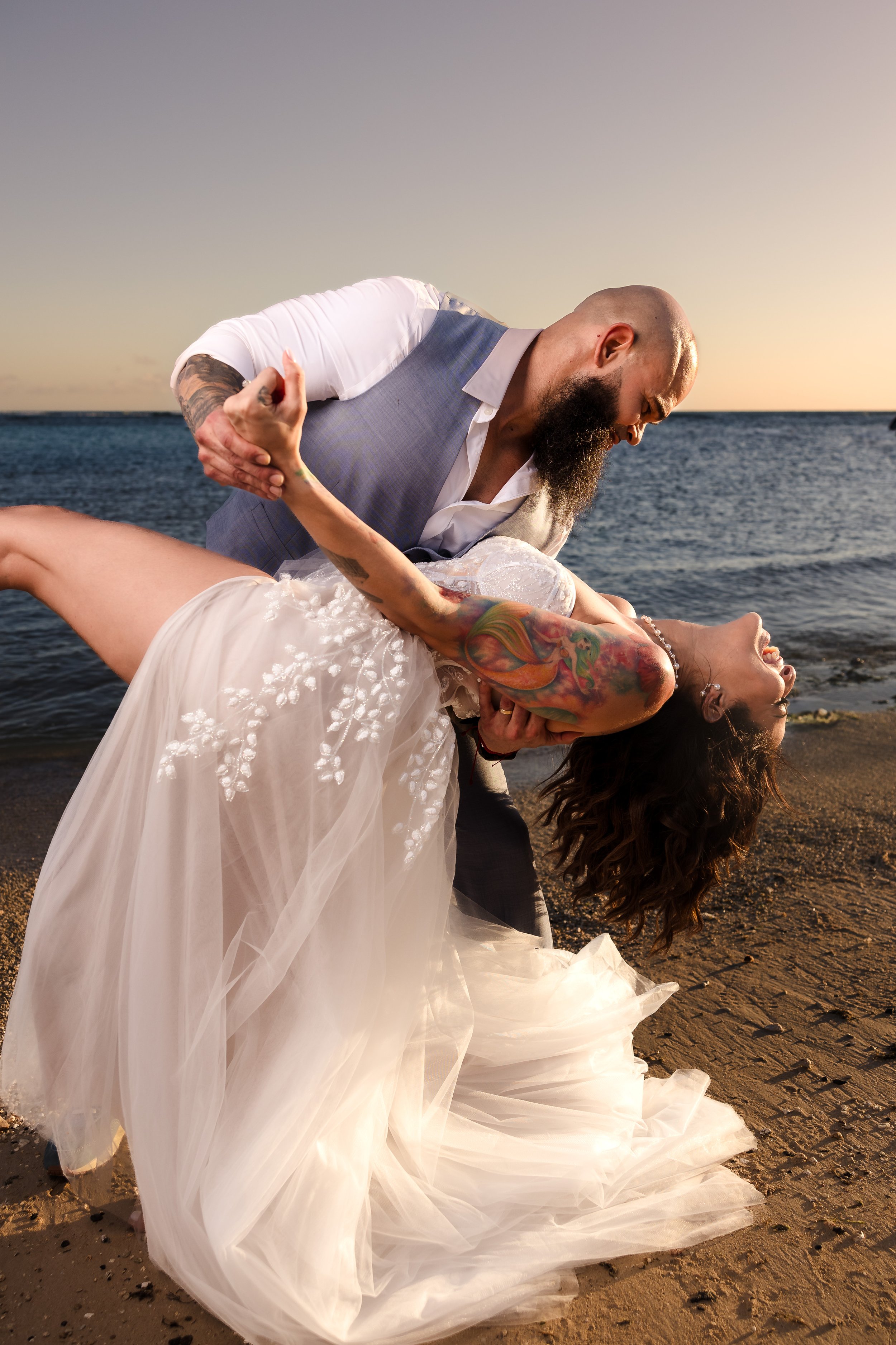 A couple is dancing on the beach during sunset, with the man dipping the woman in a romantic pose. The woman is wearing a white wedding dress with intricate embroidery, and the man is dressed in a light grey vest and white shirt. The ocean and sky ar