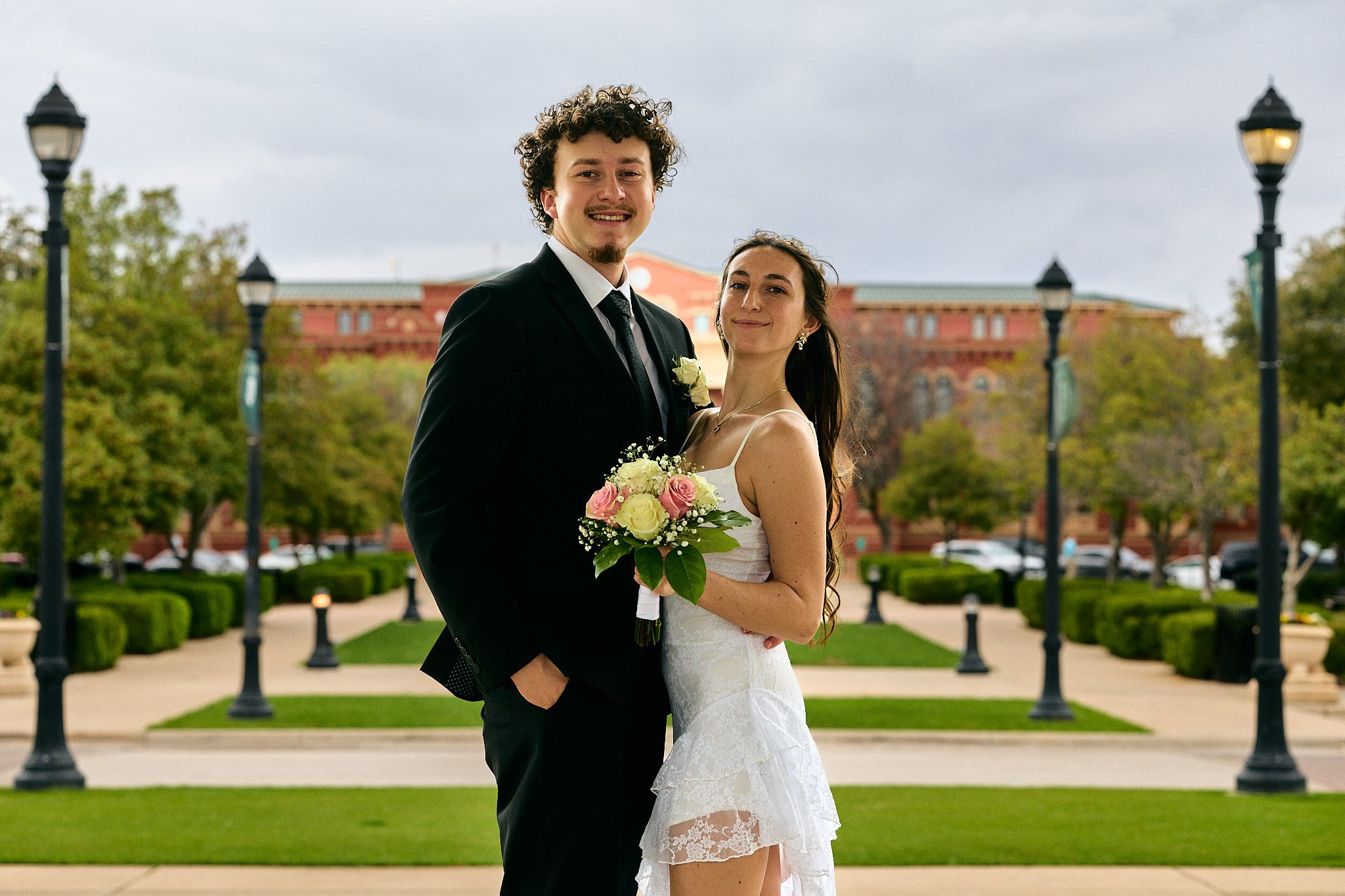 A newlywed couple standing outdoors, smiling and embracing, with the groom in a black suit and the bride in a white lace wedding dress, holding a bouquet of roses and greenery, on a pathway lined with lamp posts and hedges, with a red brick building 
