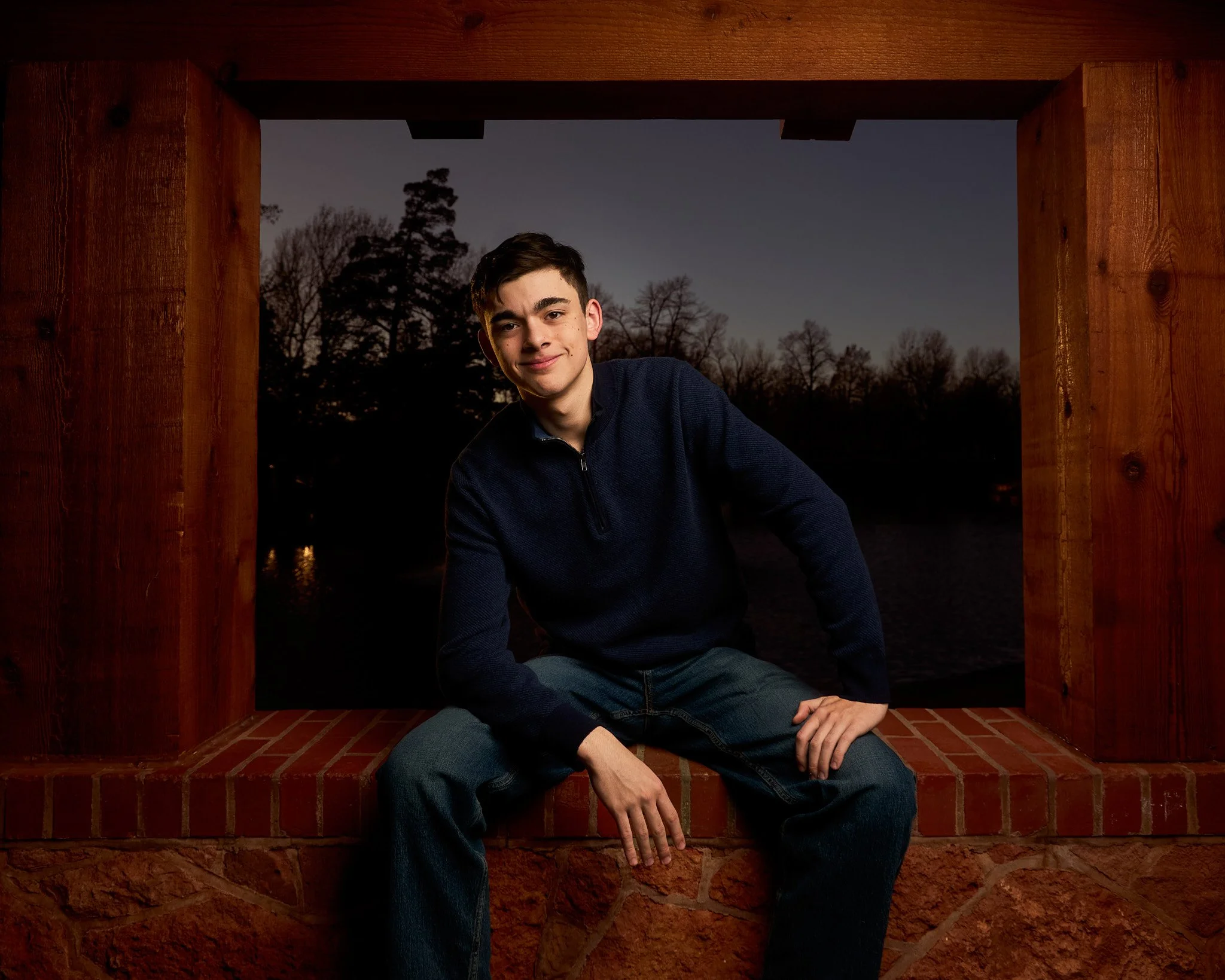 Young man sitting on a brick doorstep outdoors during dusk, framed by wooden structure, with trees in the background.