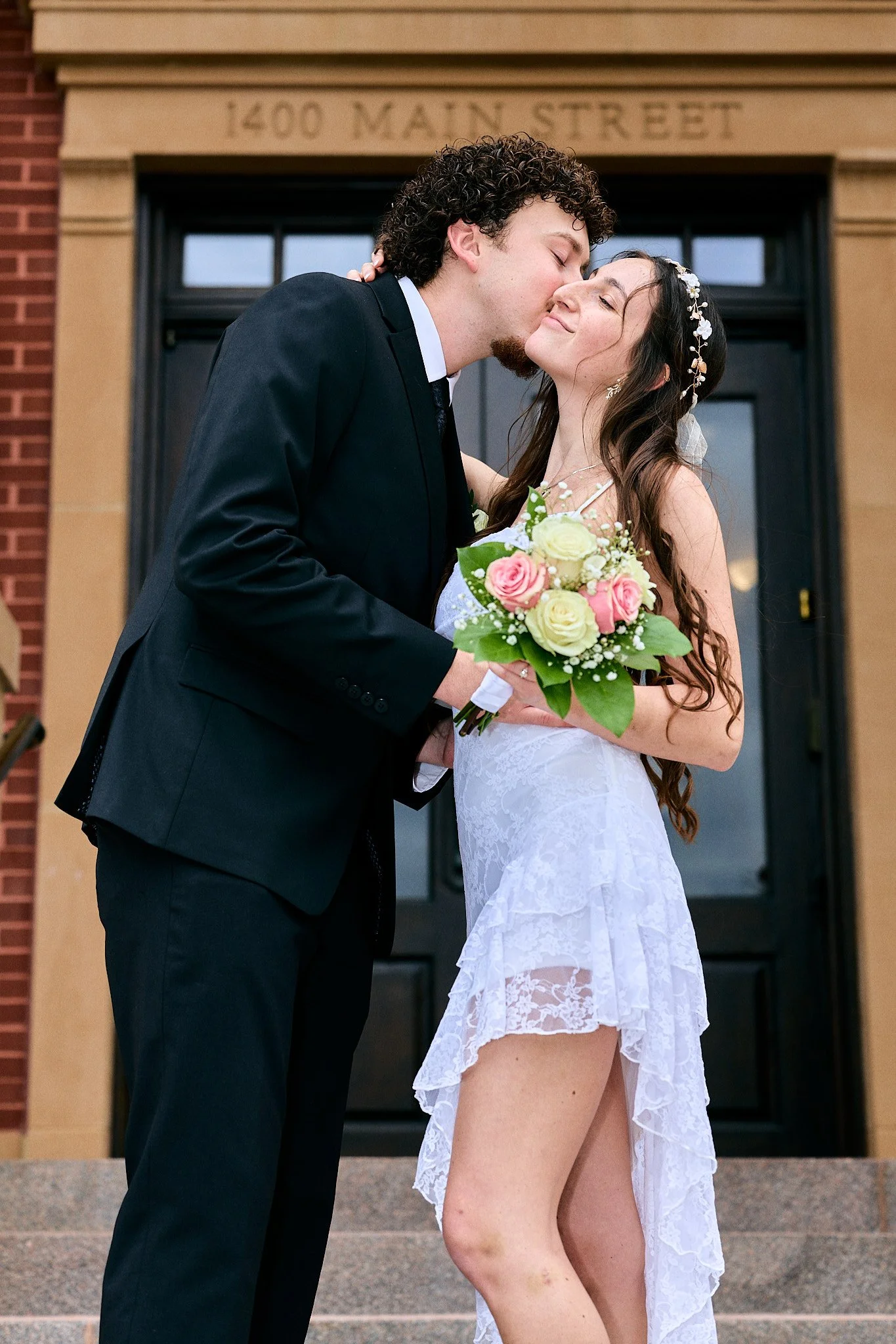 A newly married couple sharing a kiss outside a building at 1400 Main Street. The bride holds a bouquet of pink and white roses and wears a white lace dress with an asymmetrical hem, while the groom wears a black suit and tie.