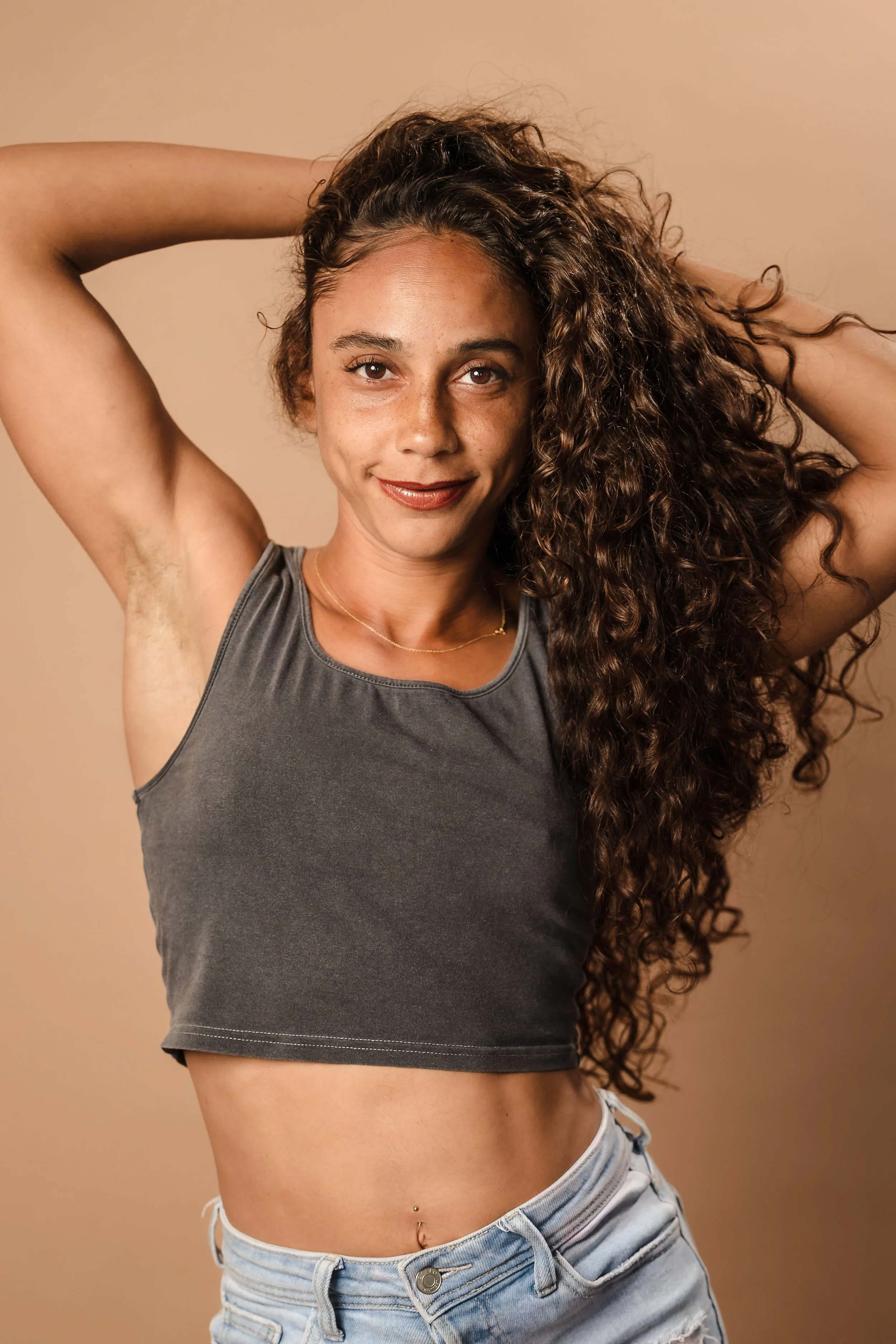 A woman with curly brown hair, posing with her arms raised above her head, wearing a grey sleeveless top and light denim shorts against a beige background.