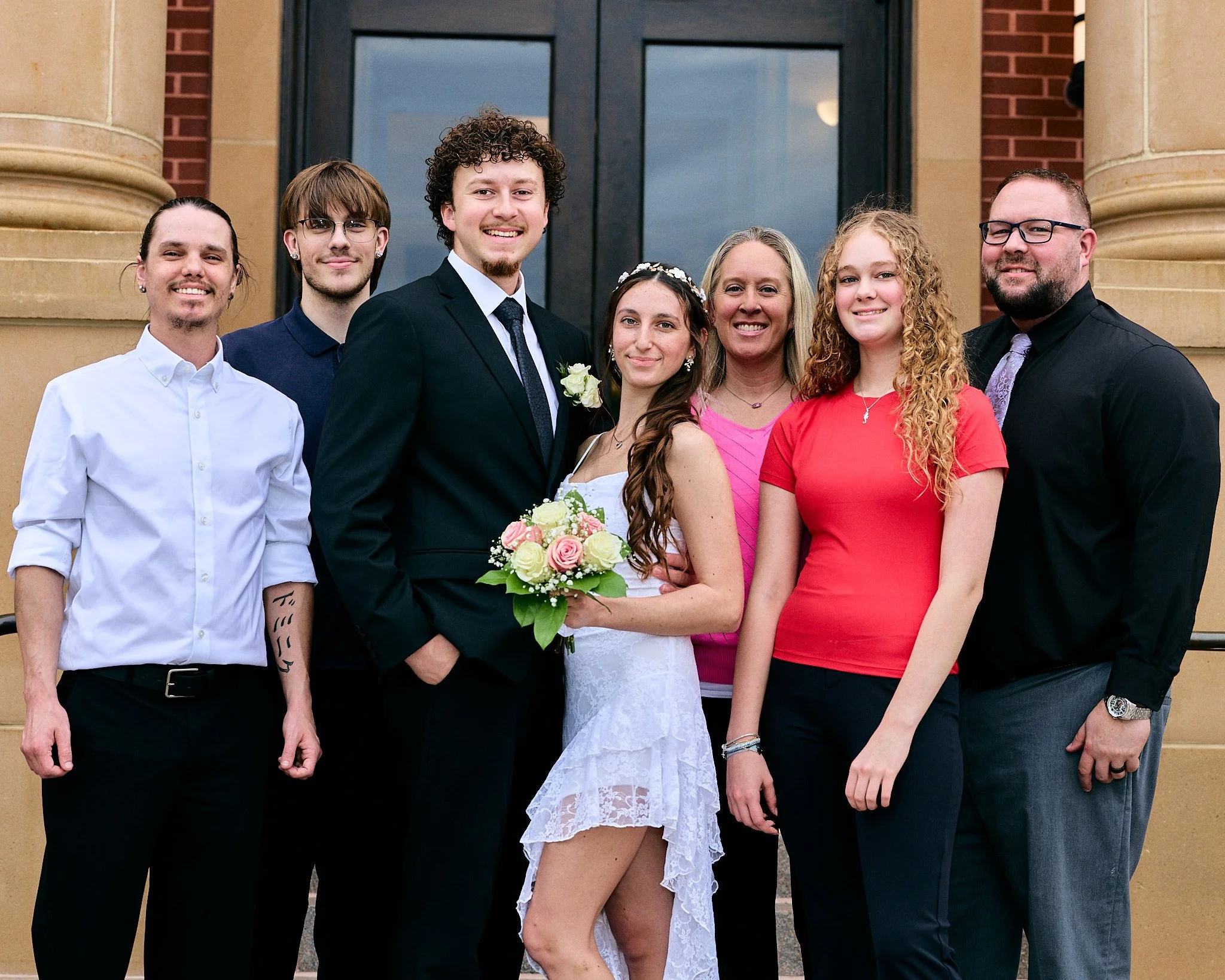 A group of seven smiling people standing in front of a building with large black doors and tan and red brick walls. Two women are wearing dresses, one holding a bouquet of flowers, and men are dressed in suits and shirts. They appear to be at a celeb