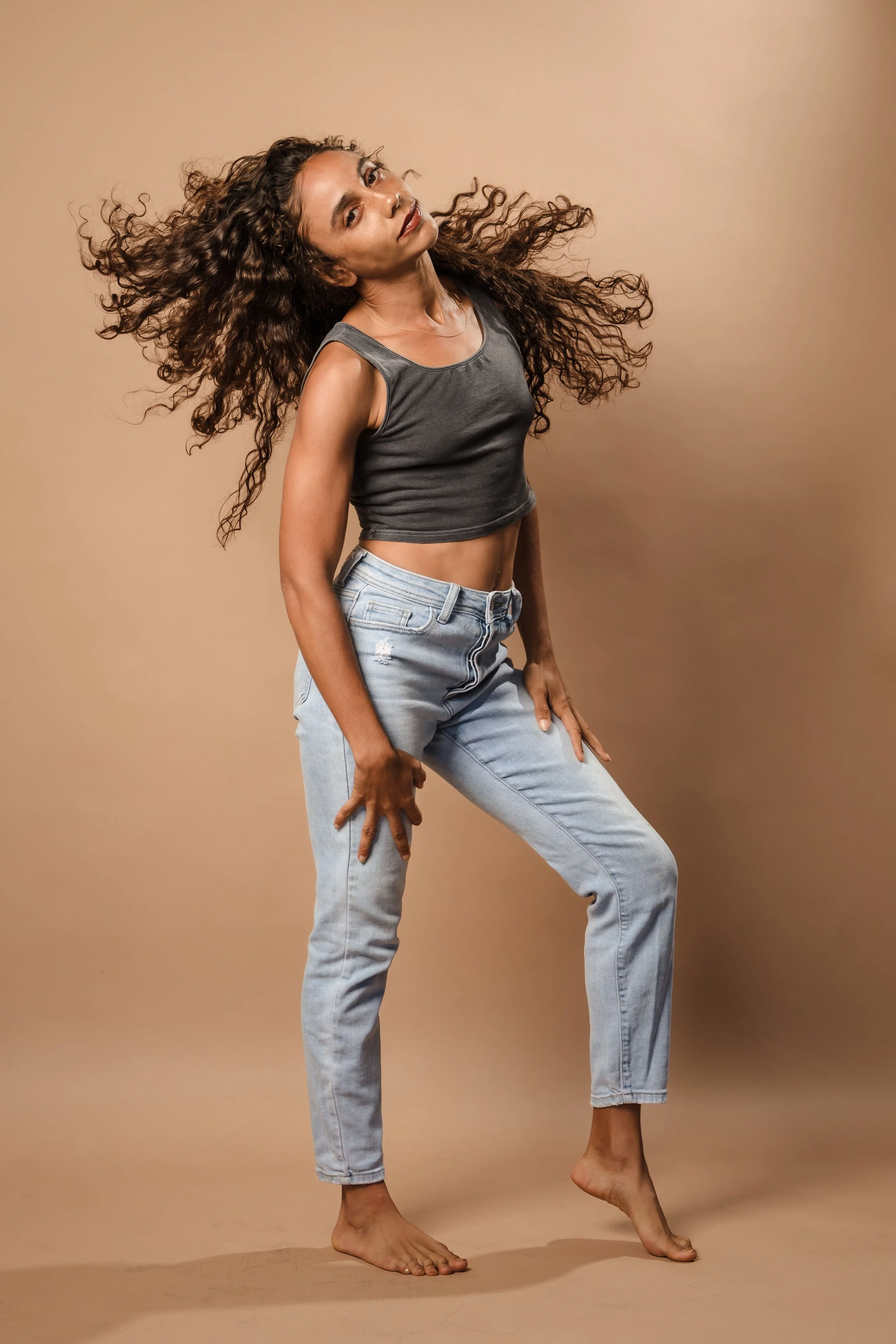 A woman with curly hair standing barefoot on a beige background, wearing a black sleeveless crop top and ripped jeans, posing with one leg bent and her moving hair.
