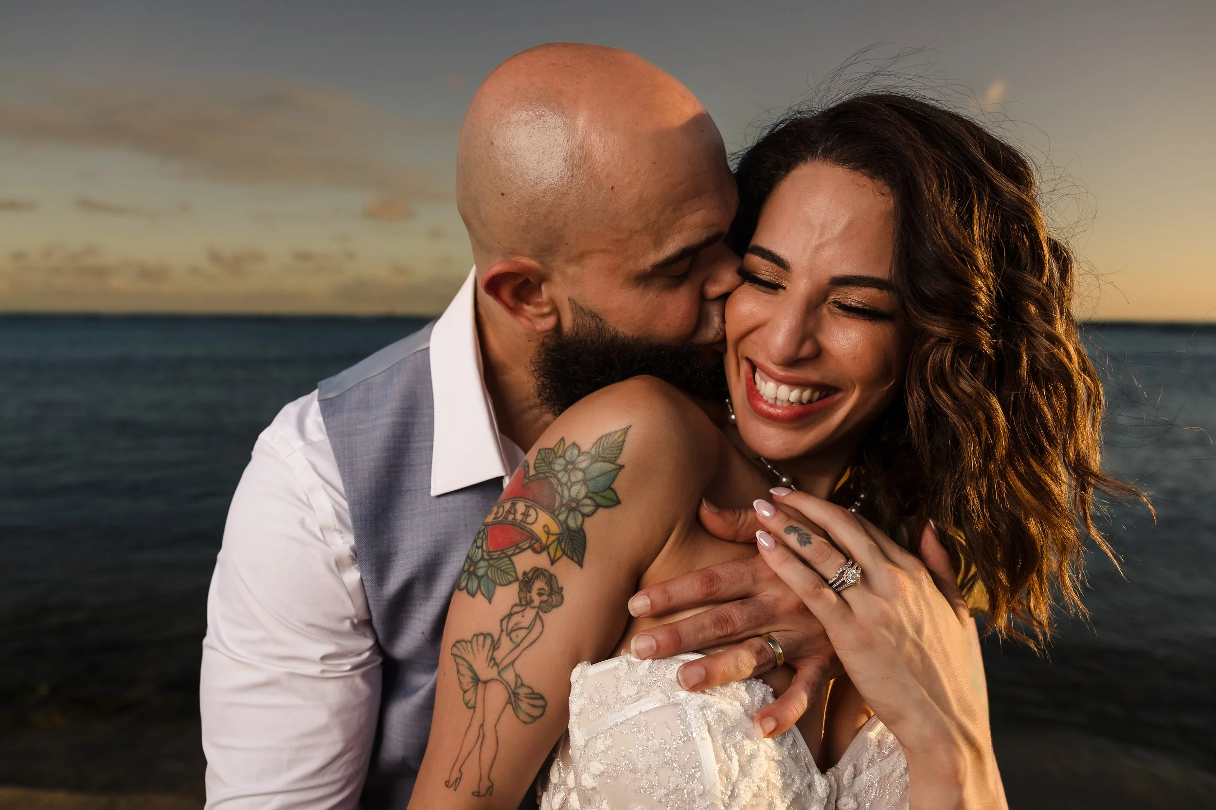 A man with a bald head and beard kisses a smiling woman with curly brown hair on the cheek, near the ocean during sunset.