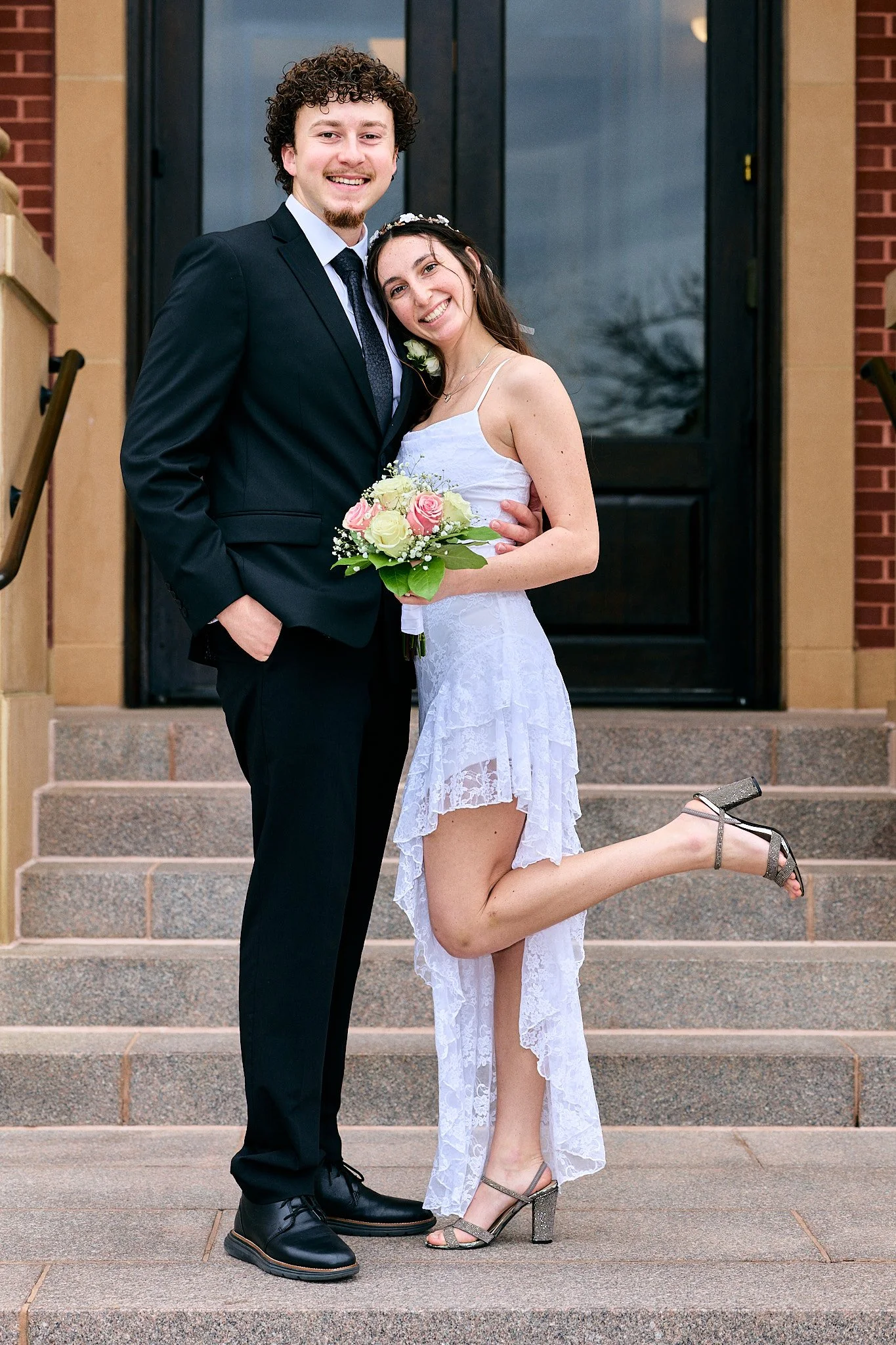 A happy couple dressed in wedding attire standing on the steps in front of a dark door. The bride is crouching on one leg, holding a bouquet of pink and white roses, and wearing a white lace dress and silver heels. The groom is in a black suit and ti
