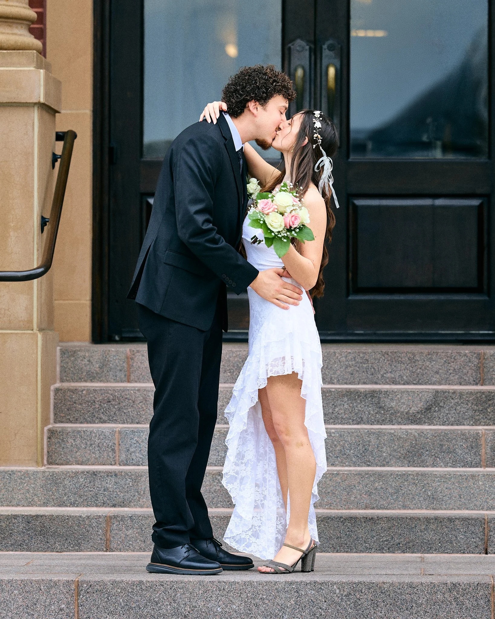 A man and woman in wedding attire share a kiss on the steps of a building. The woman is holding a bouquet of pink and white roses, and the man is wearing a dark suit.